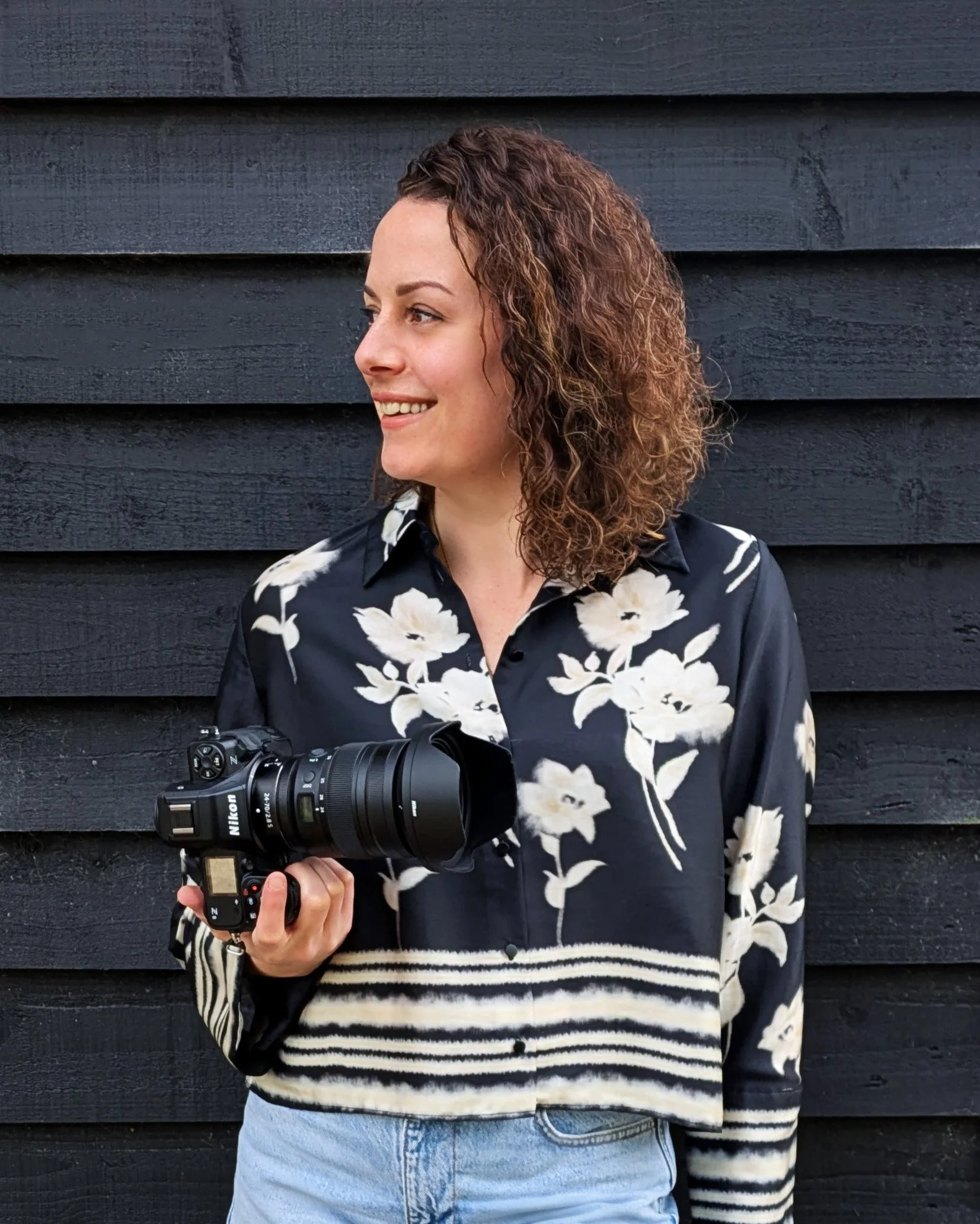 Woman with curly hair holding a camera, standing against a black wooden wall. kiss photo by steph. Wedding photographer bedfordshire based