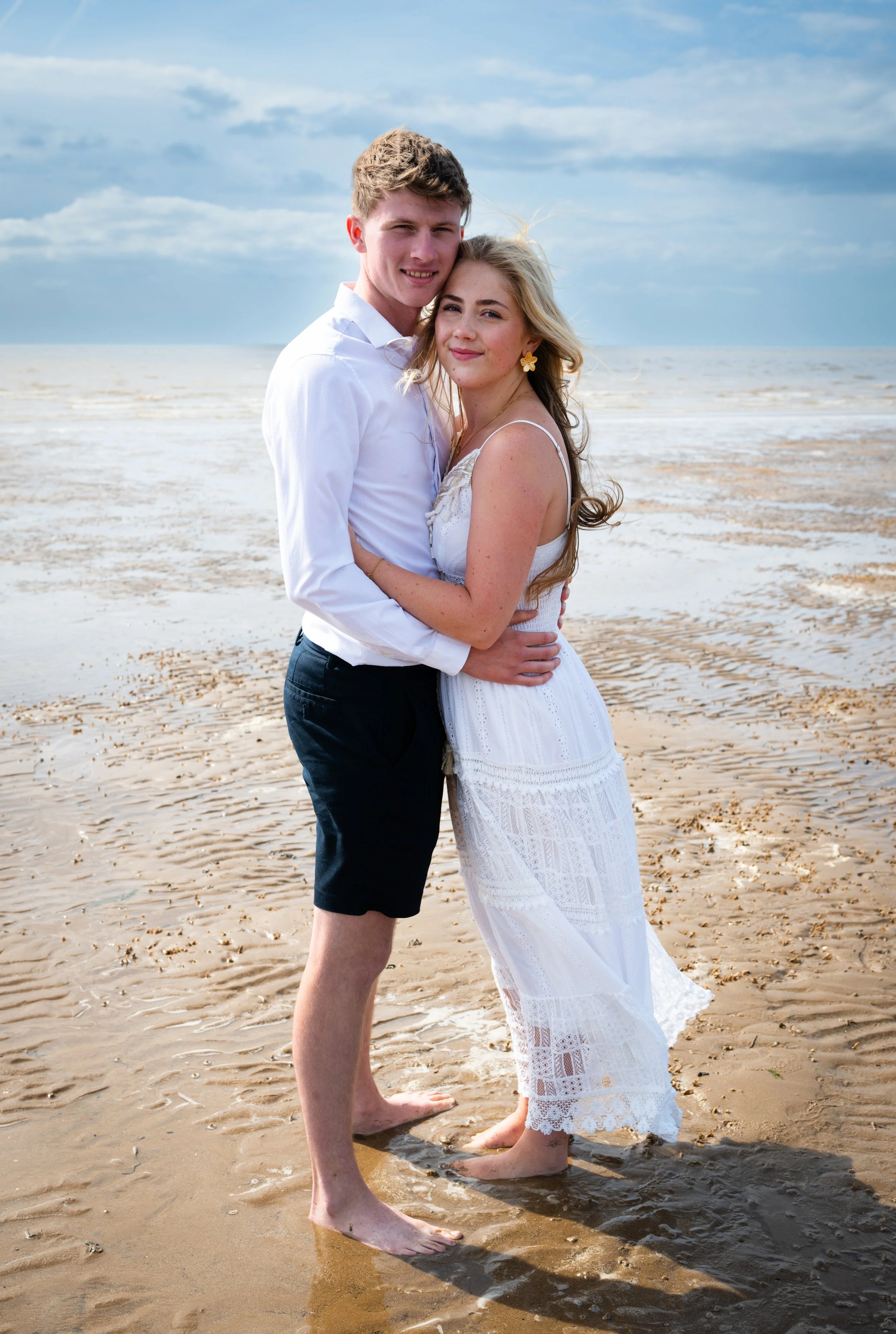 A young couple hugging on a sandy beach with waves and a cloudy sky in the background.