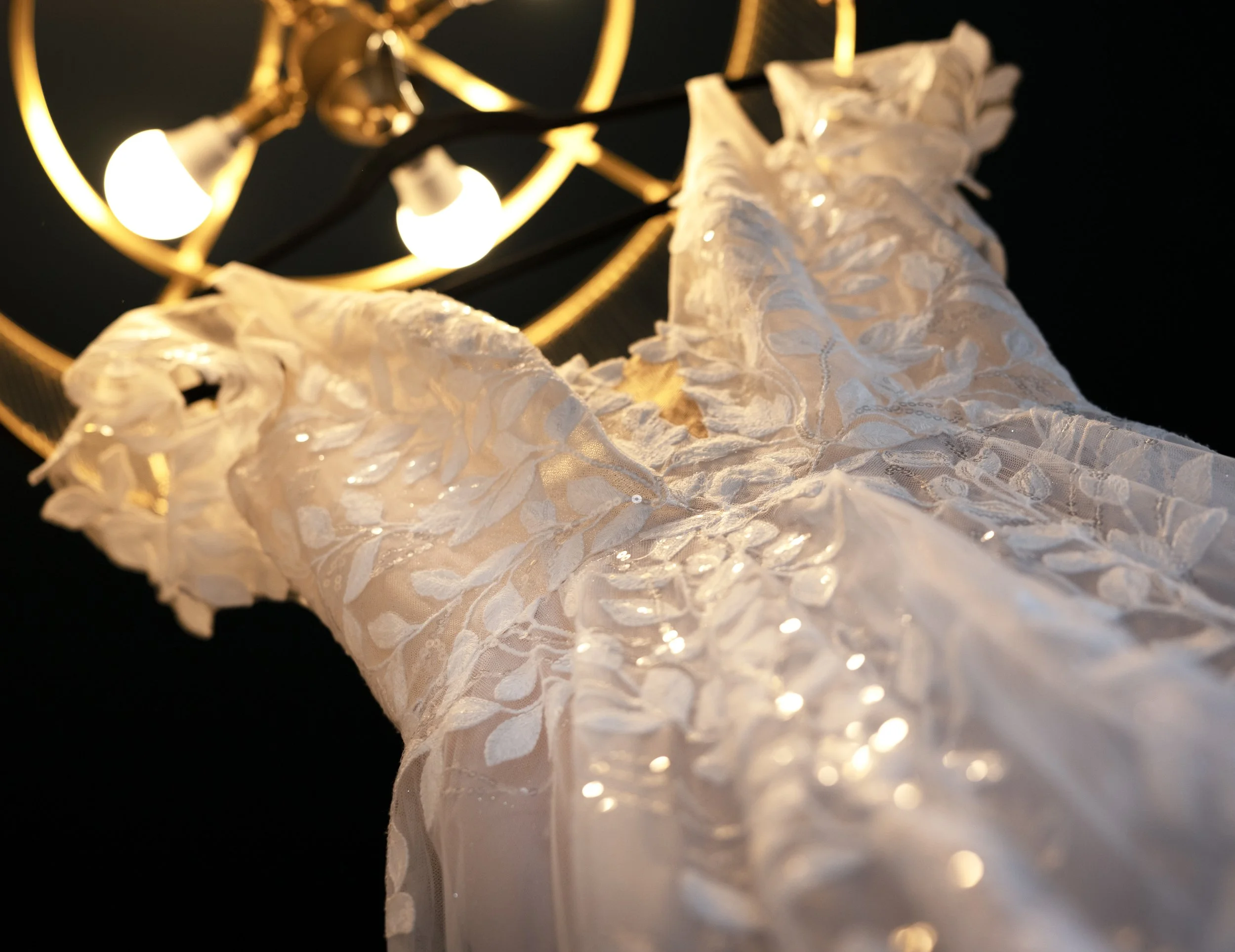 Close-up of a white, embroidered wedding dress hanging below a gold chandelier with exposed light bulbs. Wedding dress. Wedding gown.