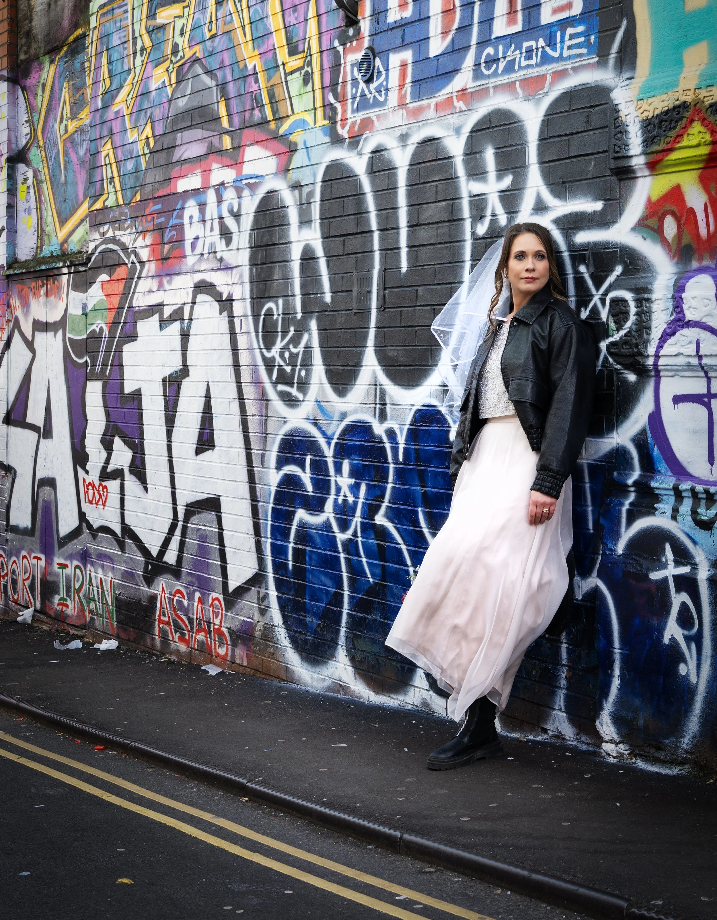 A young bride in a black leather jacket, white dress, and black boots standing against a graffiti-covered brick wall. Bristol Stokes Croft 