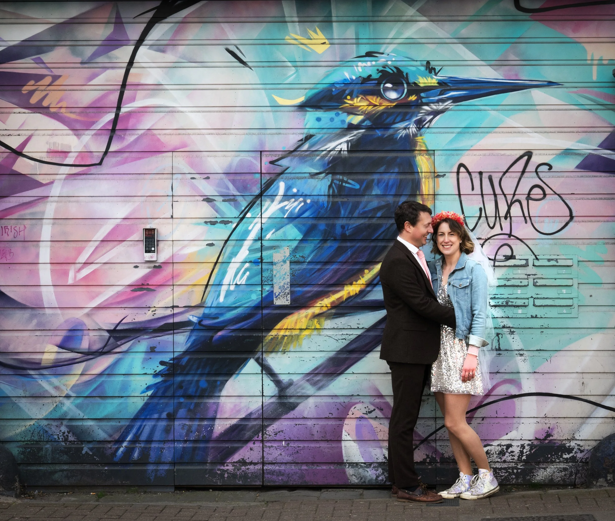 A bridal couple stands in front of colorful graffiti mural of a large blue and yellow bird on a wall. Bristol North Street