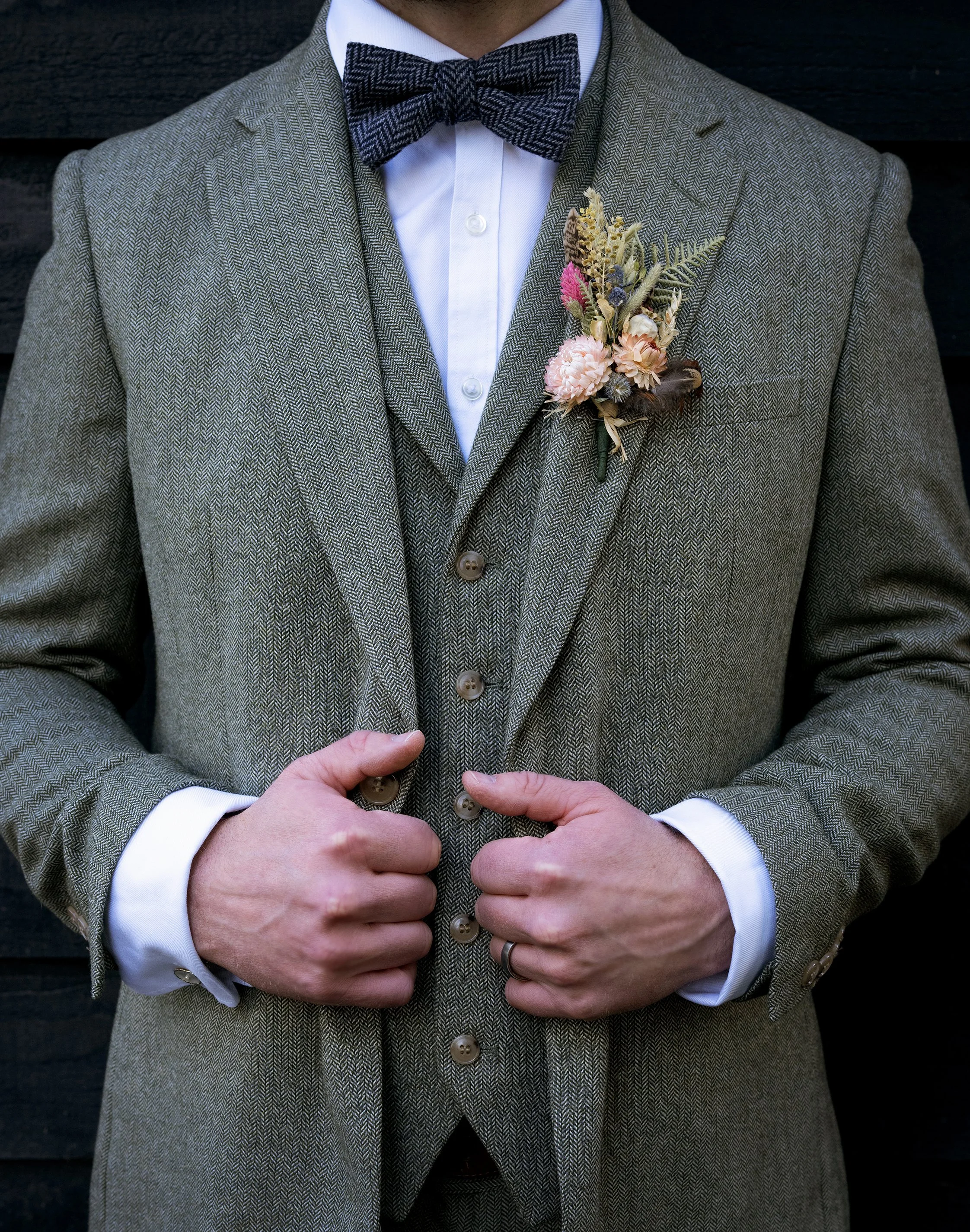 Close-up of a groom in a grey tweed suit with a white dress shirt and a bow tie, holding the front of his jacket with both hands, showing a floral boutonniere on his left lapel.