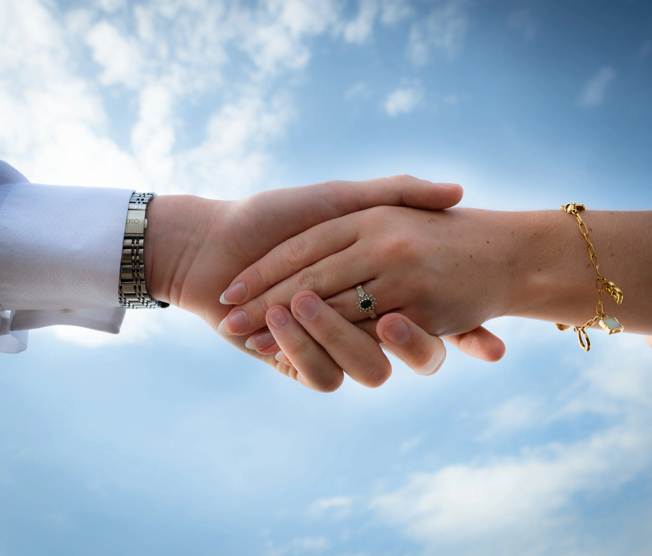 Two people holding hands against a blue sky with clouds, one person wearing a silver watch and a bracelet, the other wearing a ring and a gold bracelet.