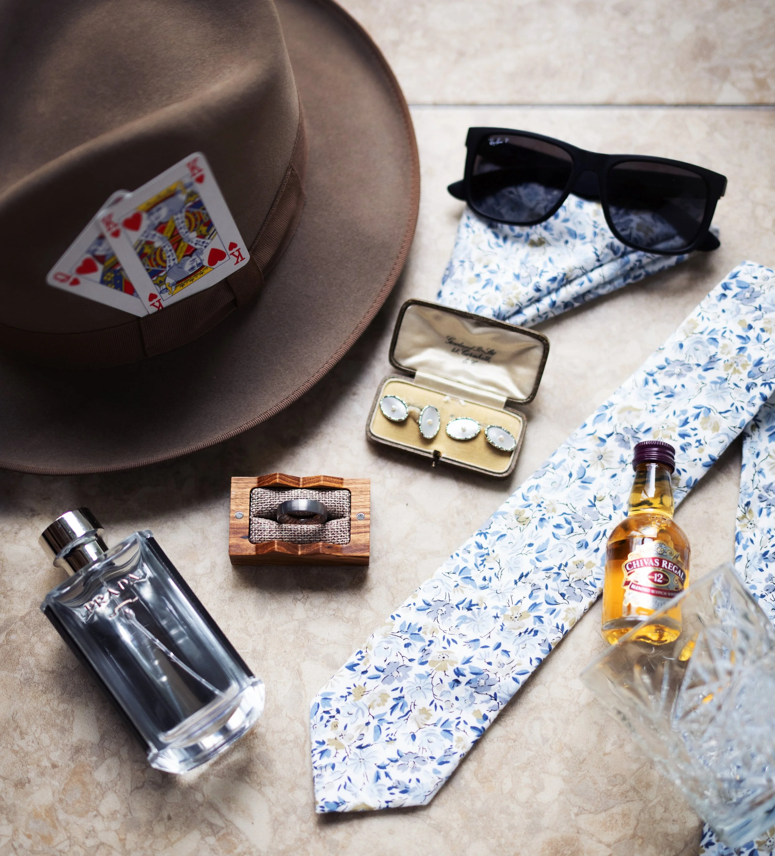 Groom wedding accessories, including a brown hat with playing cards, black sunglasses with a floral cloth underneath, a small tin case with cufflinks, a wooden ring box, a bottle of cologne, a bottle of Chivas Regal 12-year-old whiskey. Flatlay.