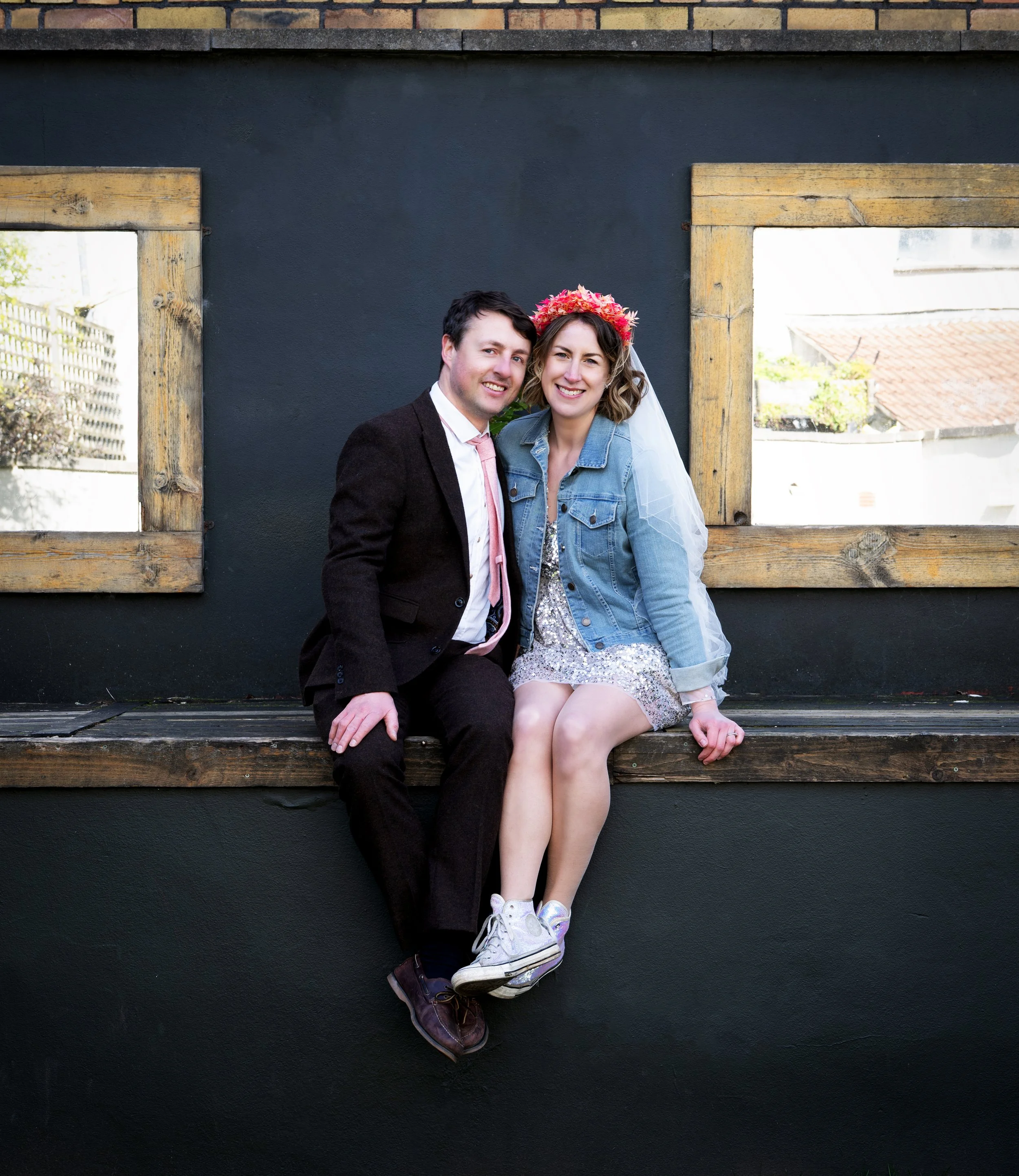 A wedding couple sitting on a wooden bench in front of a black wall with two windows, one of which has a view of a rooftop and trees visible through the glass. Bristol pub wedding.