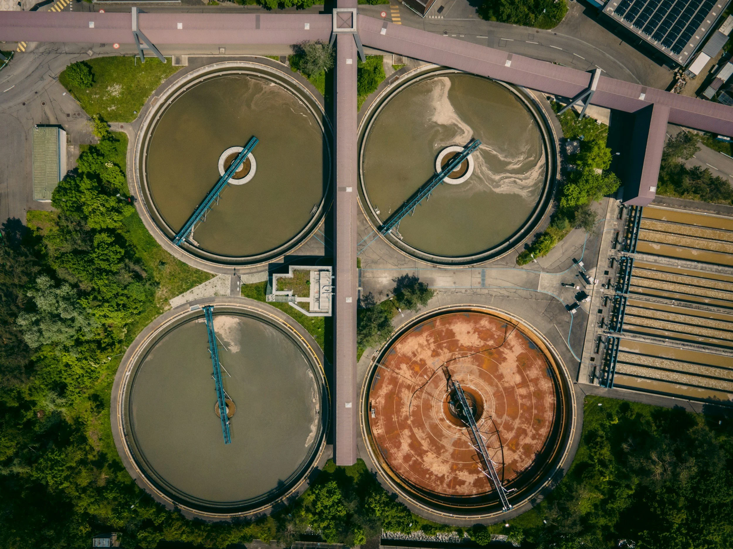 Aerial view of four large circular water treatment tanks, three filled with water and one with rust-colored residue, connected by walkways and surrounded by trees and industrial buildings.