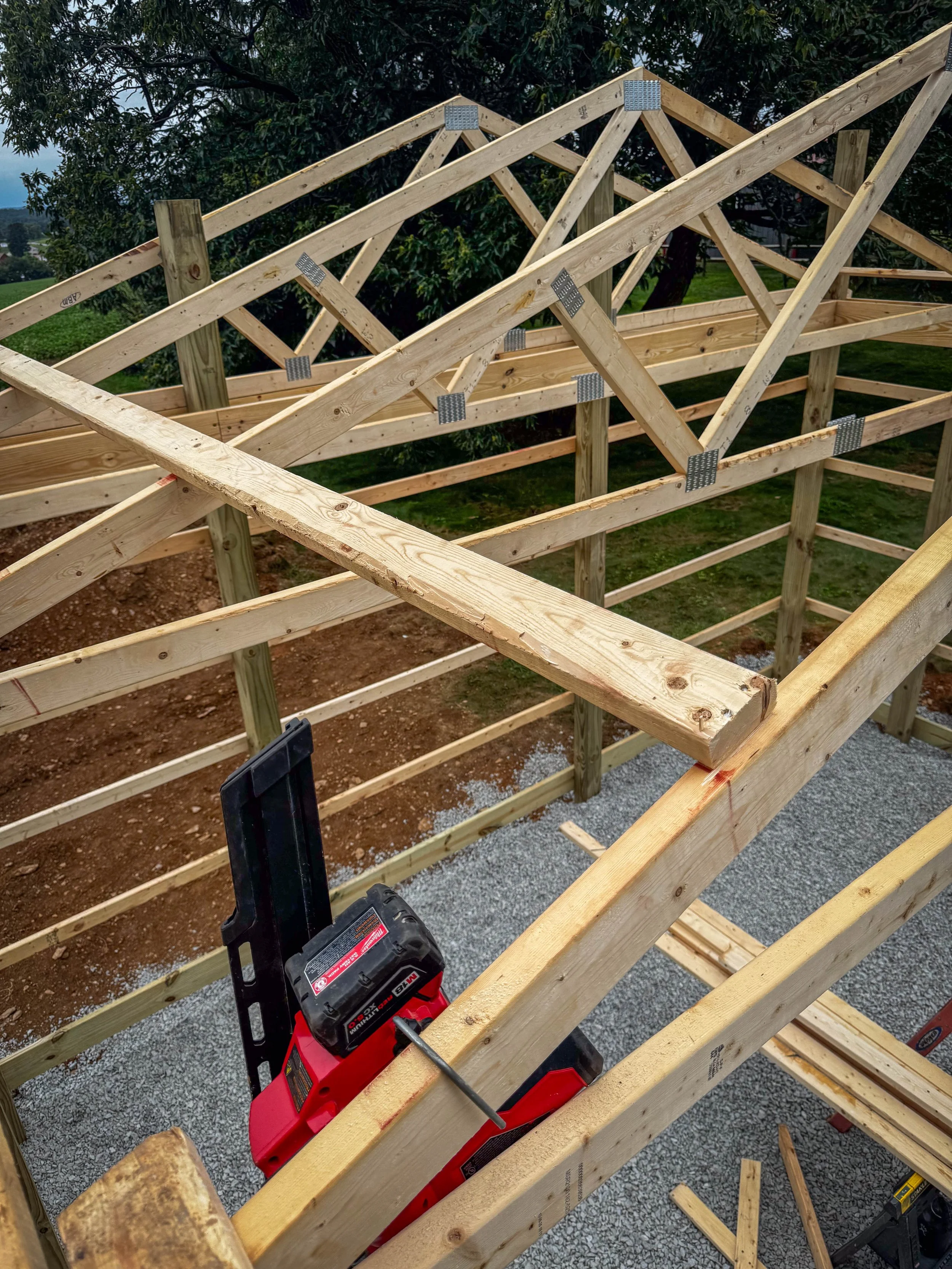 Construction site with wooden framing and a red power tool on gravel ground, with trees and greenery in the background.