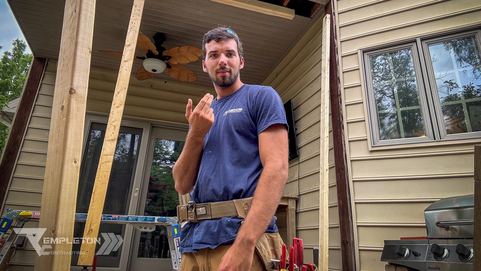 A man wearing a blue t-shirt and a construction tool belt stands in front of a house undergoing renovation. The house has beige siding, windows, and a partially built deck with wooden beams and a ceiling fan. Construction tools are visible in the foreground.