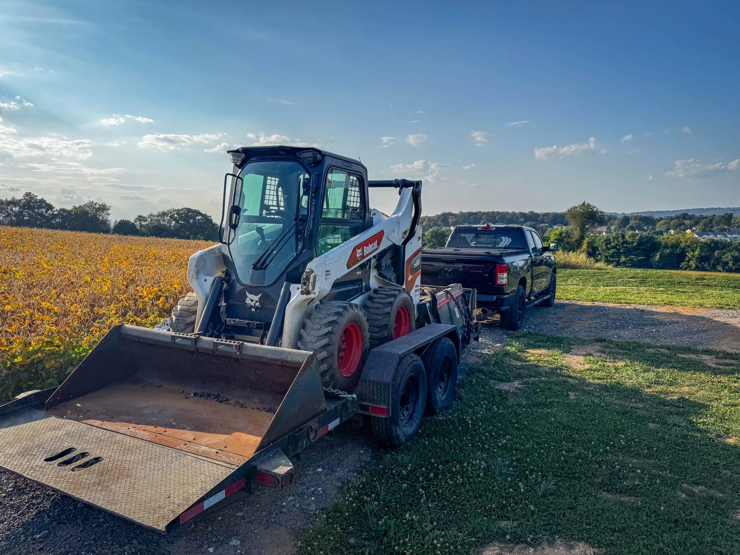 A Bobcat skid-steer loader on a trailer hitched to a black pickup truck in a rural, open field with a clear blue sky and some clouds.