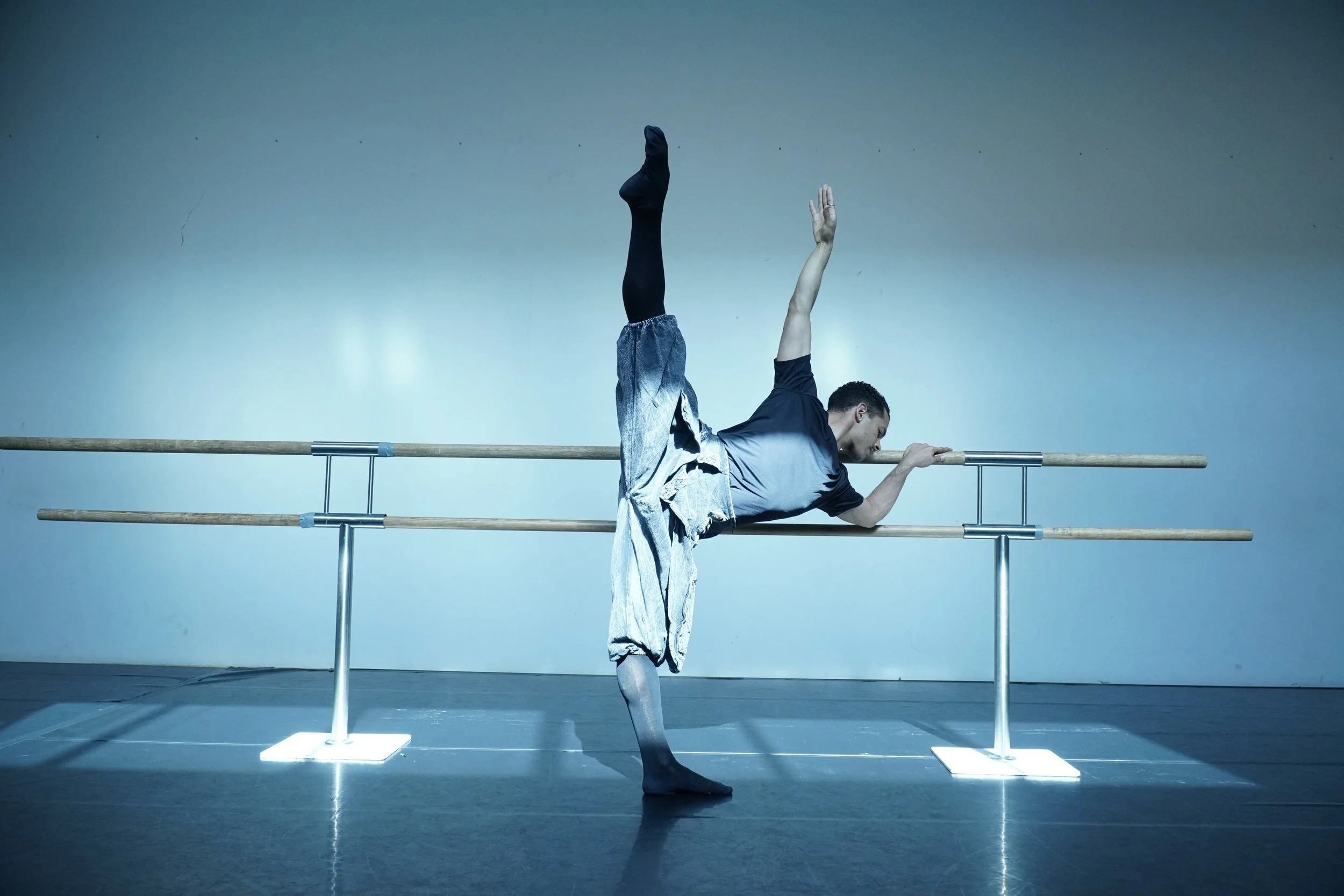 A male ballet dancer practicing at a ballet barre, balancing on one foot with the other leg raised high and his arm extended for balance.