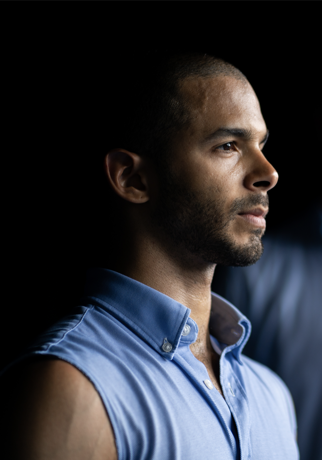 A man with short hair and a beard wearing a sleeveless blue collared shirt, looking to the right in profile against a dark background.