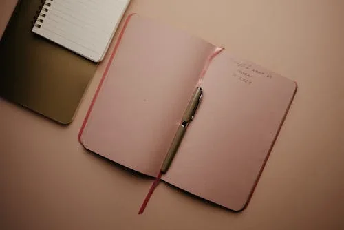 Open pink notebook with handwritten notes and a pen, partially visible nearby notebooks.
