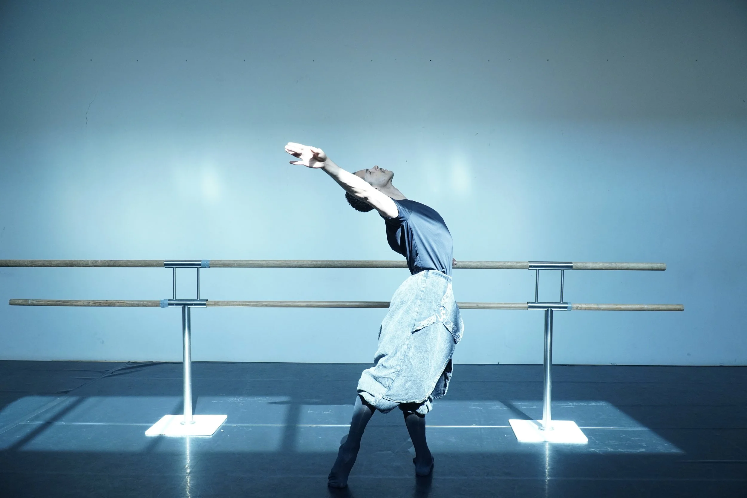 A ballet dancer practicing in front of a mirror with ballet bars, wearing a dark blue top and loose beige pants, with arms extended and head tilted back.