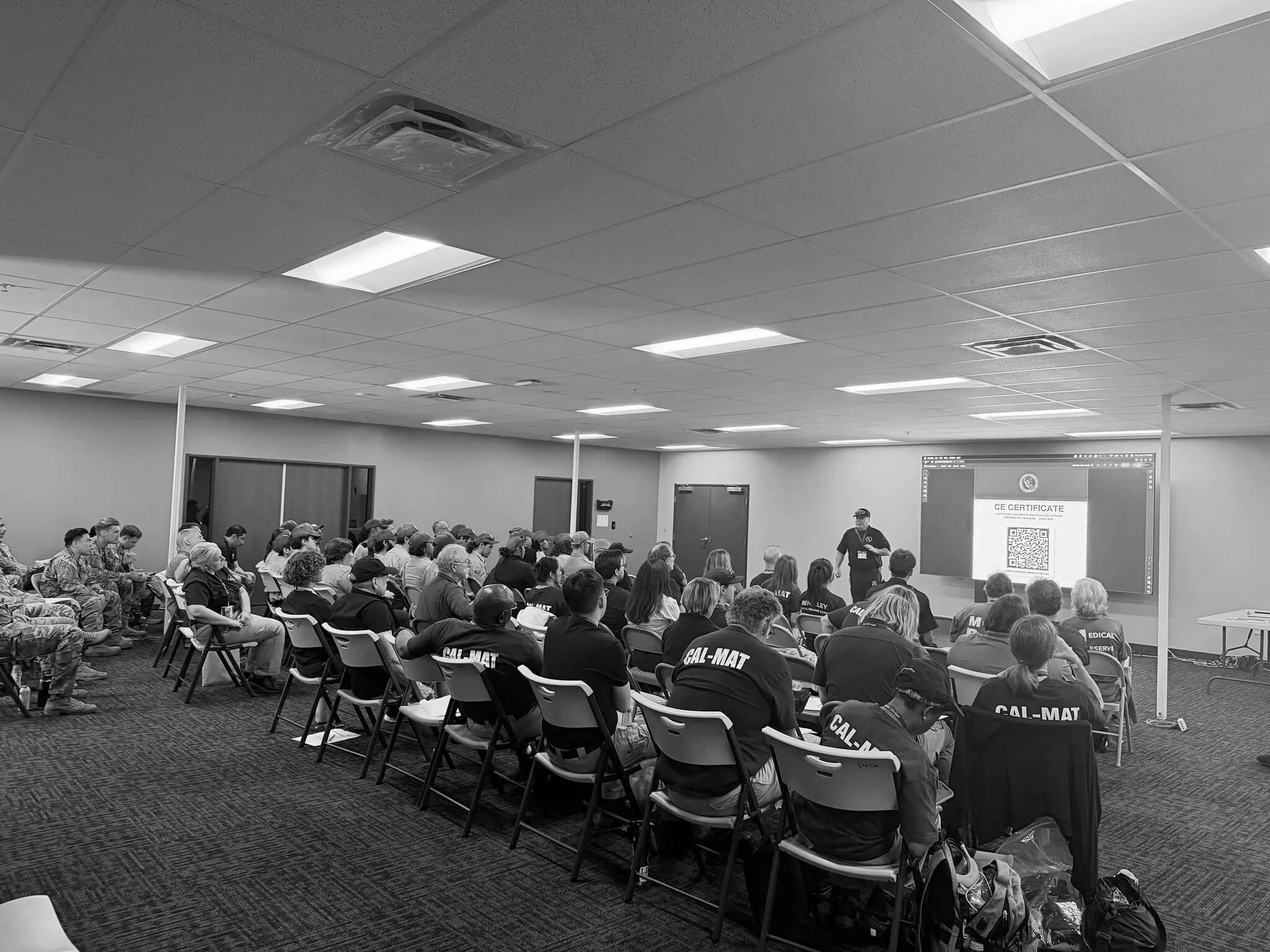 A group of uniformed individuals, some wearing CAL-MAT shirts, seated in chairs attending a presentation in a large conference room with a projector screen at the front.