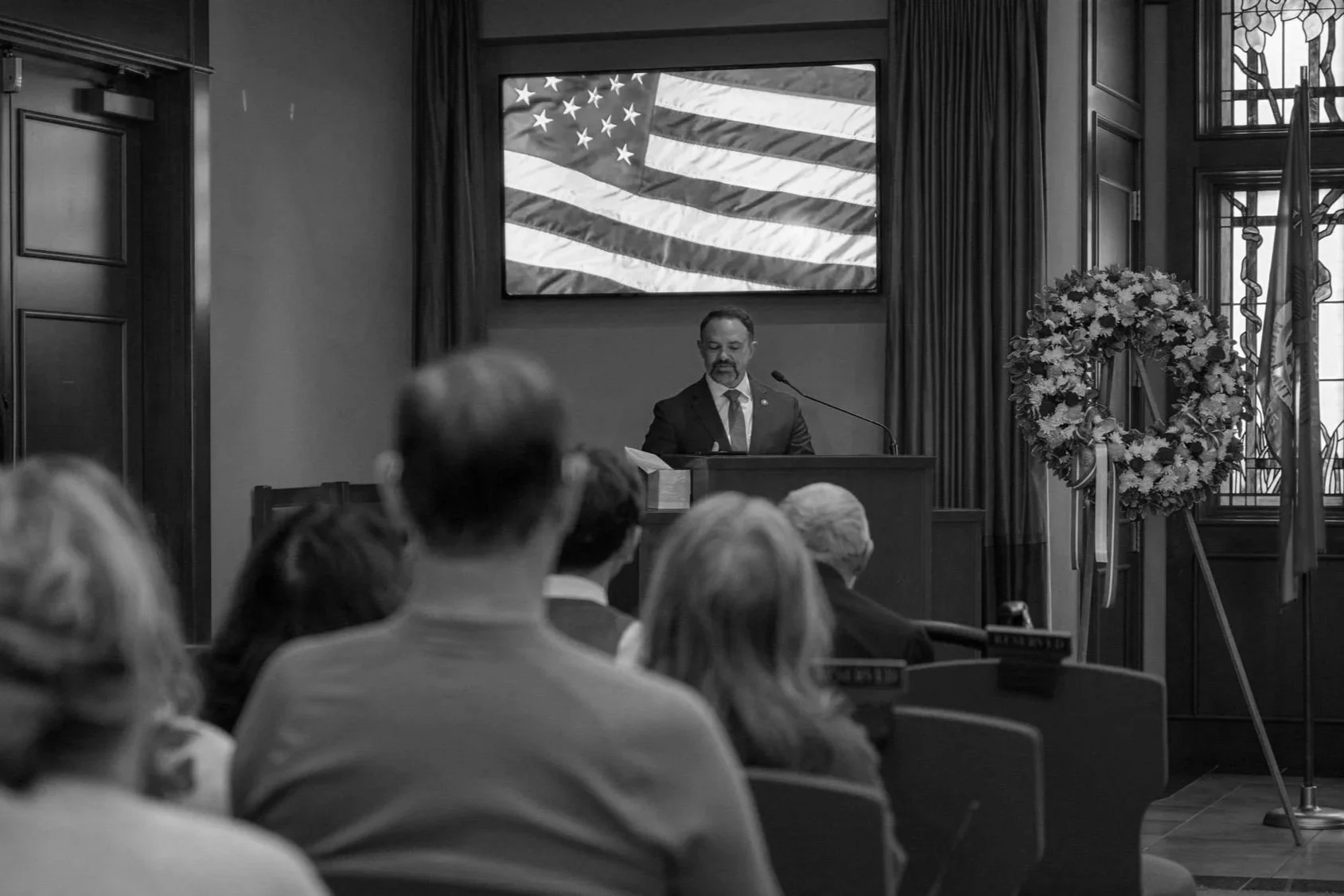 A man in a suit speaking at a podium during a formal event in a room decorated with an American flag and a floral wreath.