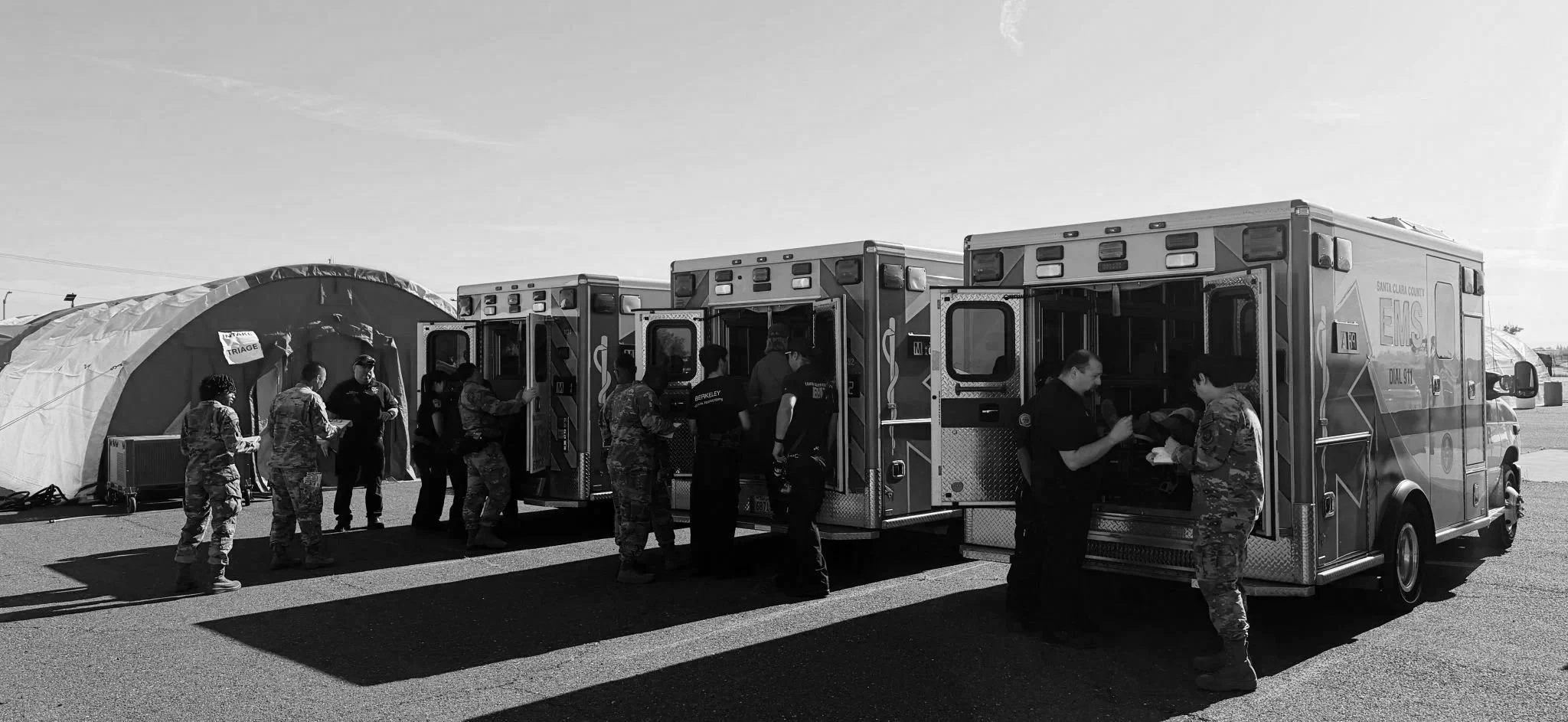 A group of people, including military personnel and emergency responders, standing outside a row of ambulance vehicles near a triage tent on a clear day.