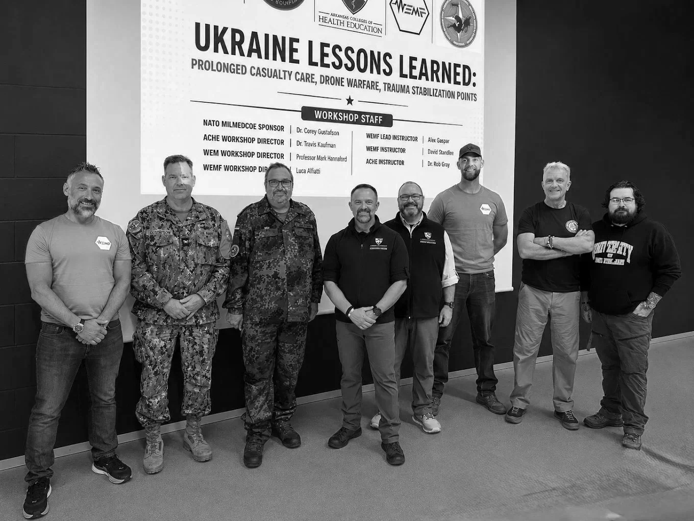 Group of eight men standing in front of a presentation screen titled 'Ukraine Lessons Learned,' with content about prolonged casualty care, drone warfare, and trauma stabilization points. The men are dressed casually, some in military or tactical clothing, posing indoors.