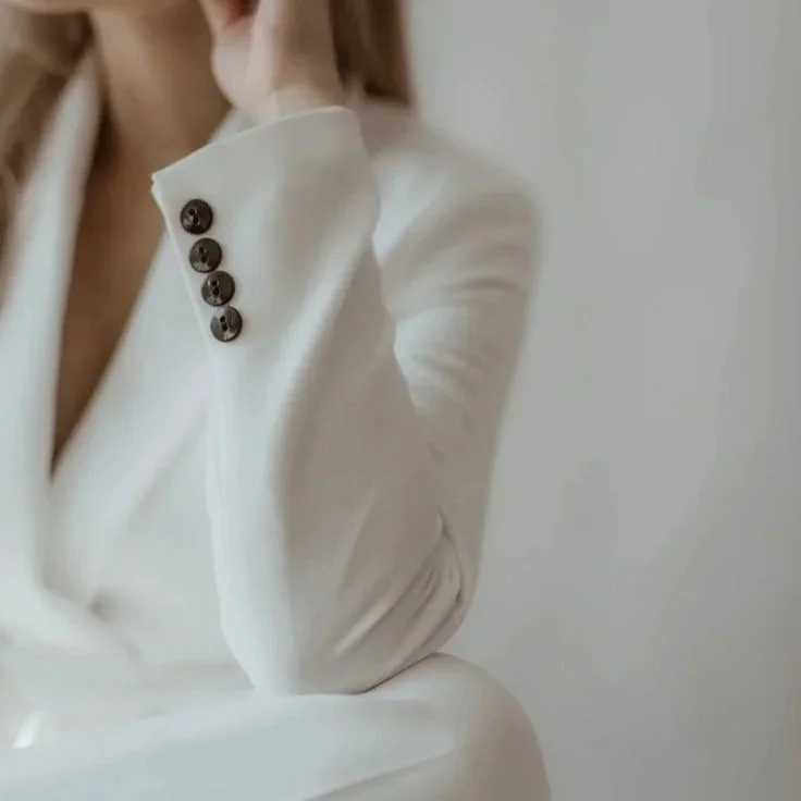 Close-up of a woman wearing a white blazer with four black buttons on the cuff, sitting with her arm resting on a surface, against a plain light-colored background