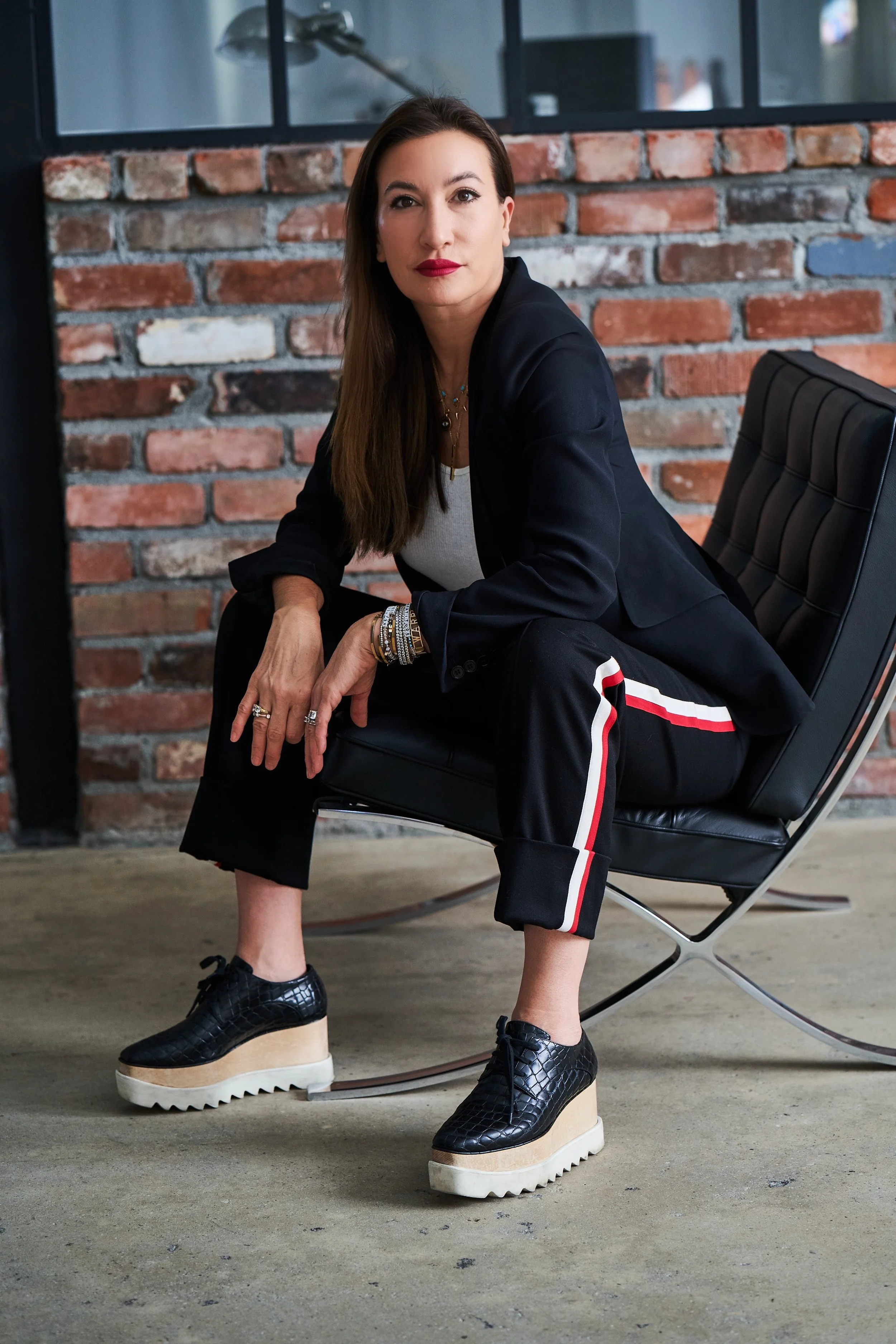 A woman with long brown hair and red lipstick sitting on a black leather chair, wearing a black blazer, white top, black pants with red and white stripe, and platform shoes with textured black upper and beige soles, in front of brick wall and industrial background.