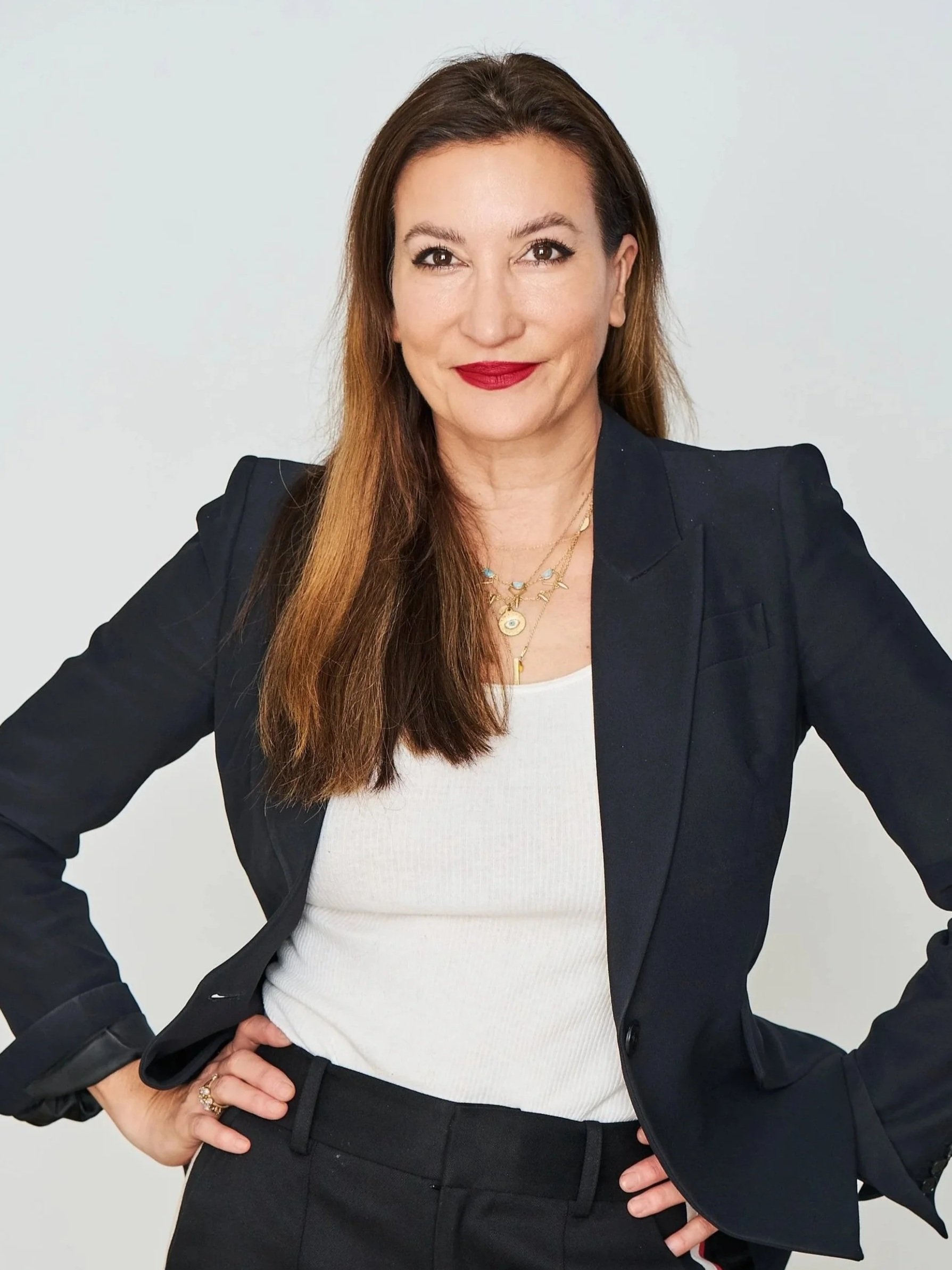 A confident woman with long brown hair, wearing a black blazer, white top, and black pants, standing with hands on hips and smiling at the camera.