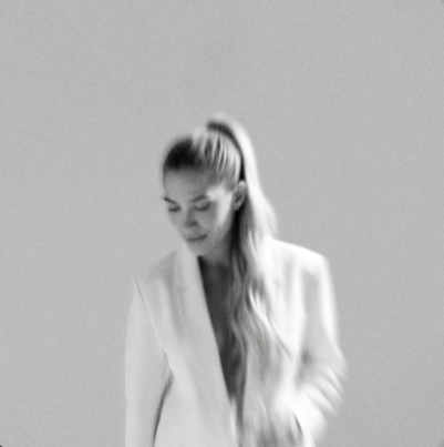 A woman with long blonde hair in a ponytail, looking down, wearing a blazer, against a plain background.