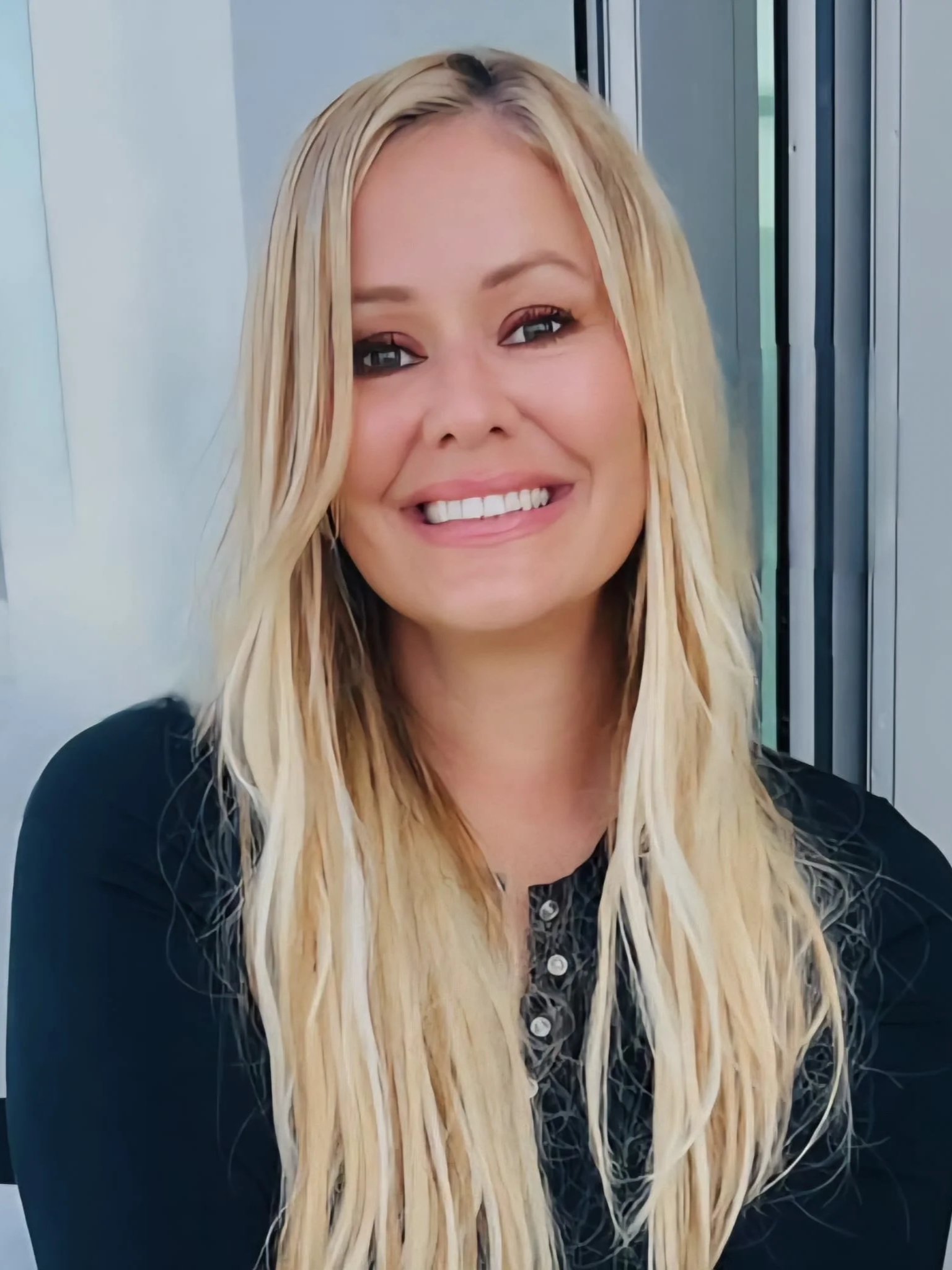 A smiling woman with long blonde hair wearing a black shirt, sitting in front of a window.