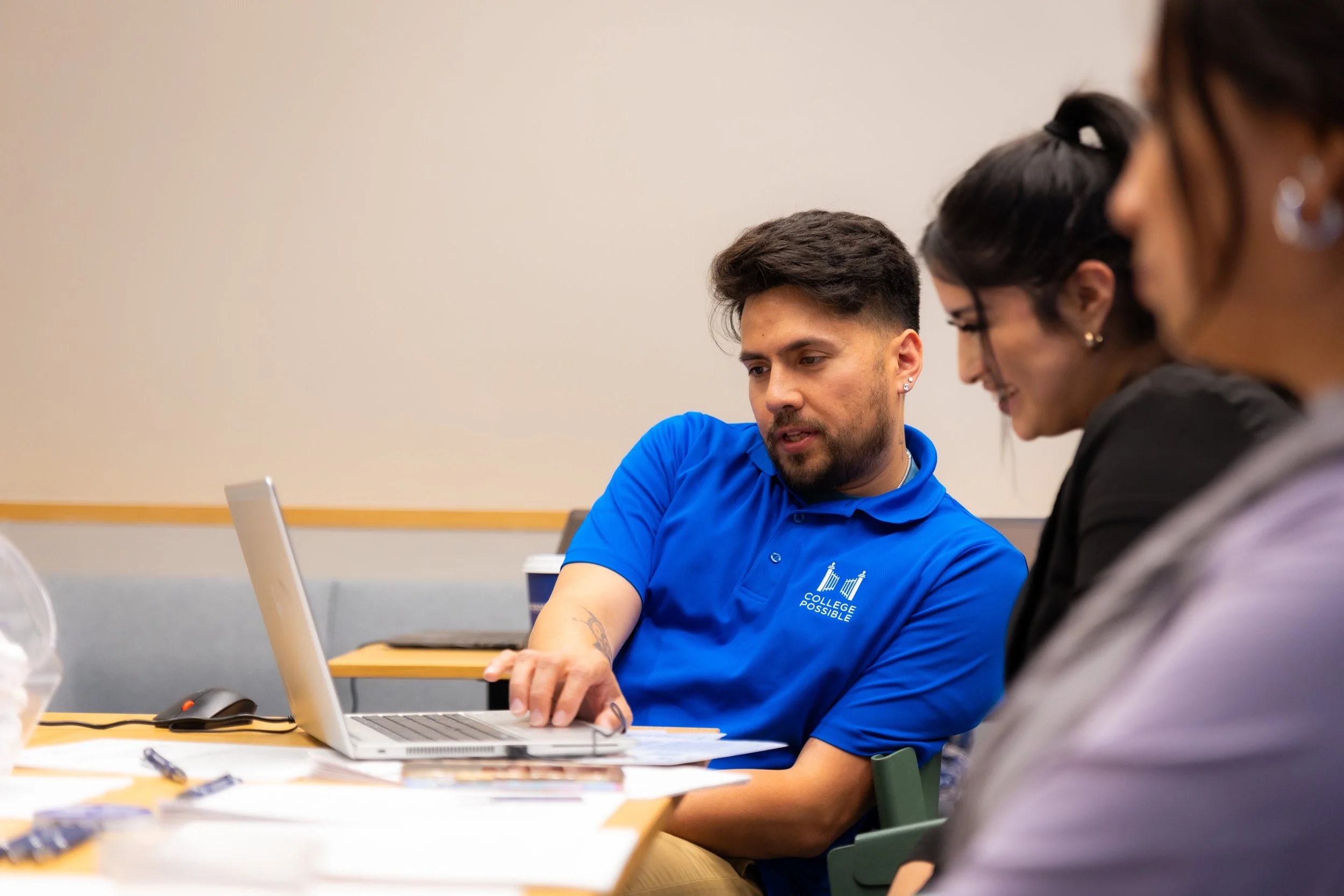 Three people sitting at a table looking at a laptop screen in a meeting or classroom setting.