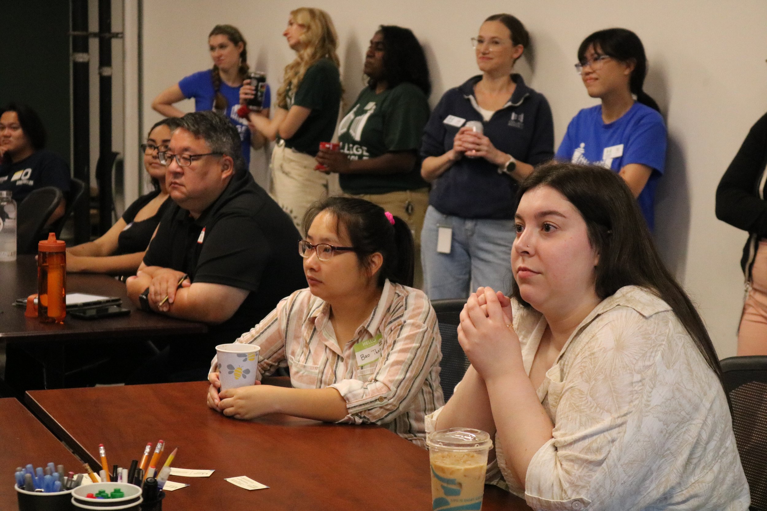 A group of diverse people listening attentively during a seminar or workshop in a conference room. Some are seated at a table with drinks and stationery, while others are standing against the wall.