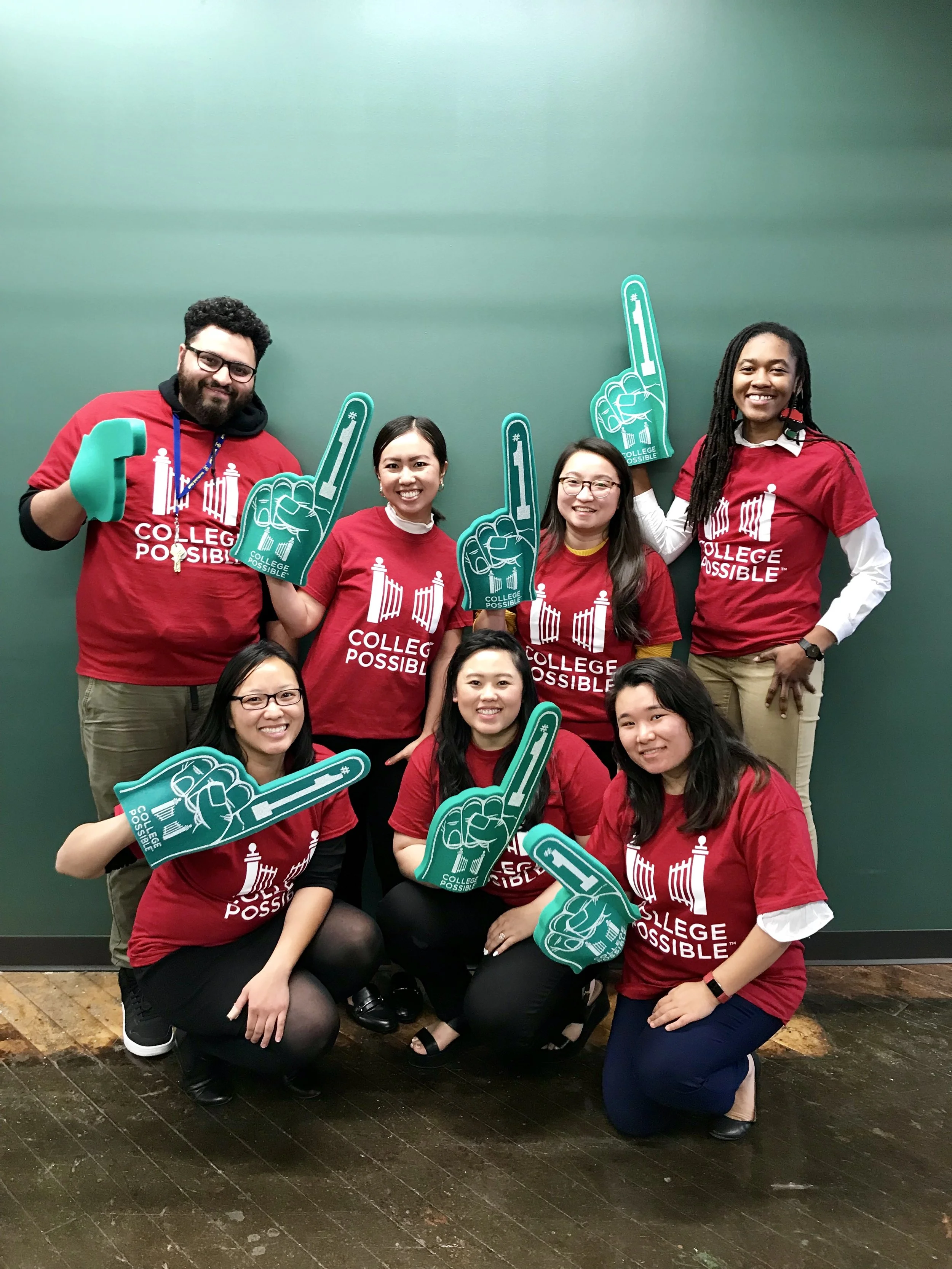 Group of seven people wearing red shirts with 'College Possible' logo, holding foam fingers, smiling, in front of a green wall.