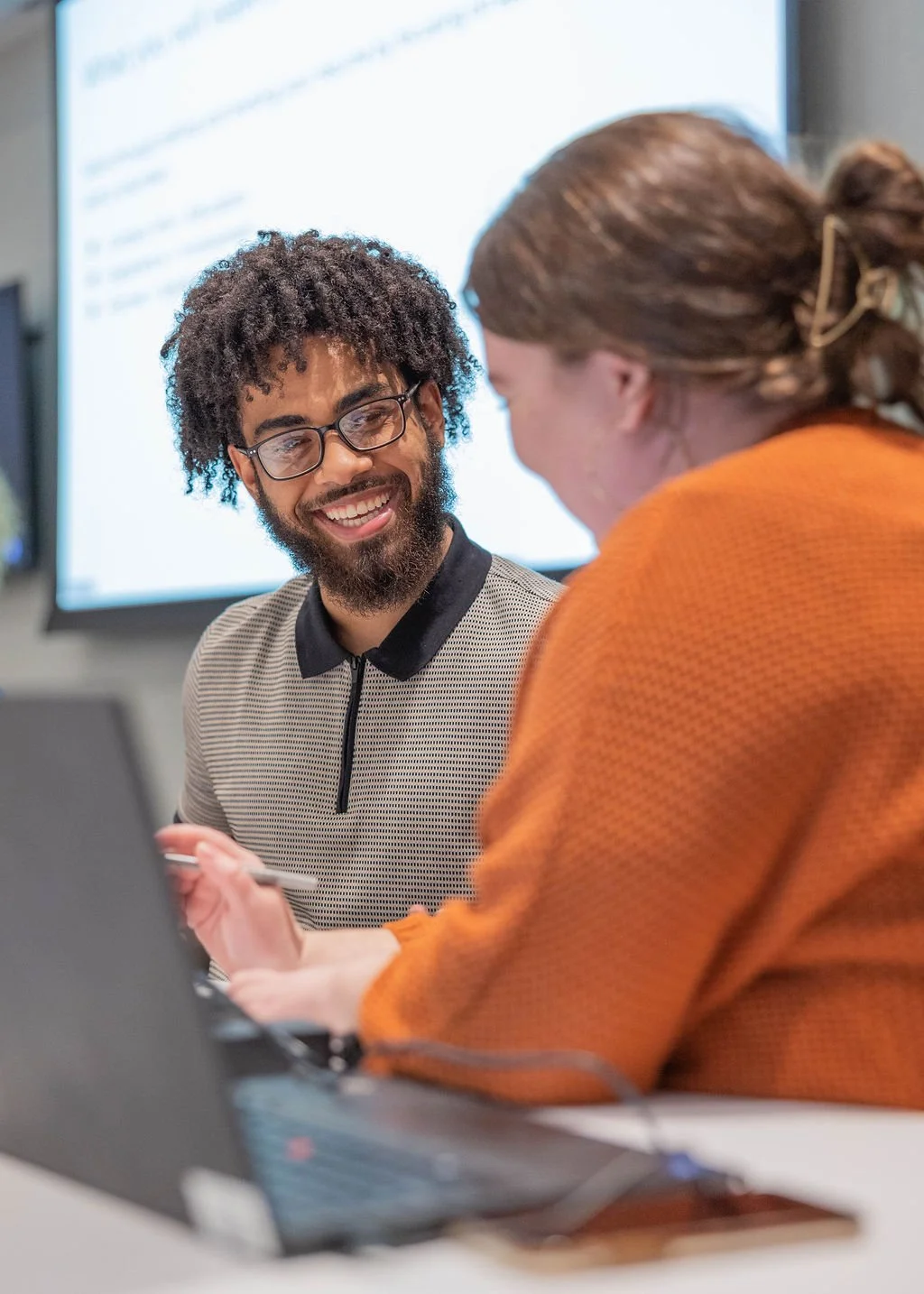 Two people engaged in a discussion in a conference room with a large screen in the background. One person has curly hair, glasses, and a beard, smiling at the other person.