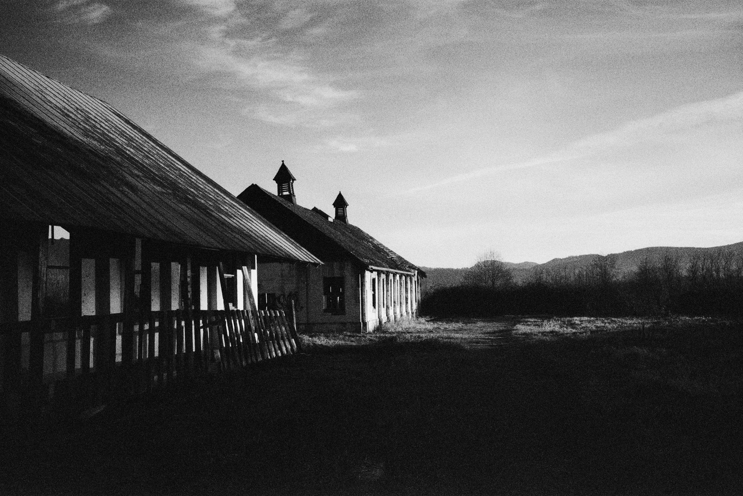 Black and white photo of an abandoned building with a slanted roof and a row of small towers on top, situated in a rural landscape with mountains in the background and a clear sky.