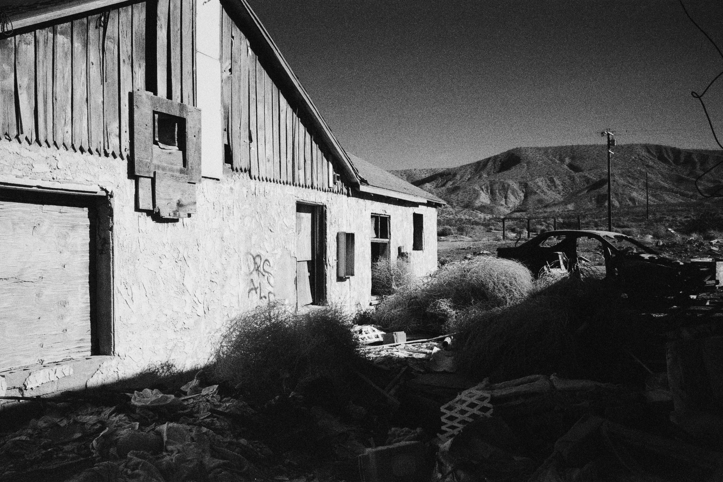 An abandoned house with boarded-up windows and a damaged car in the yard, set against a backdrop of mountains under a clear sky.