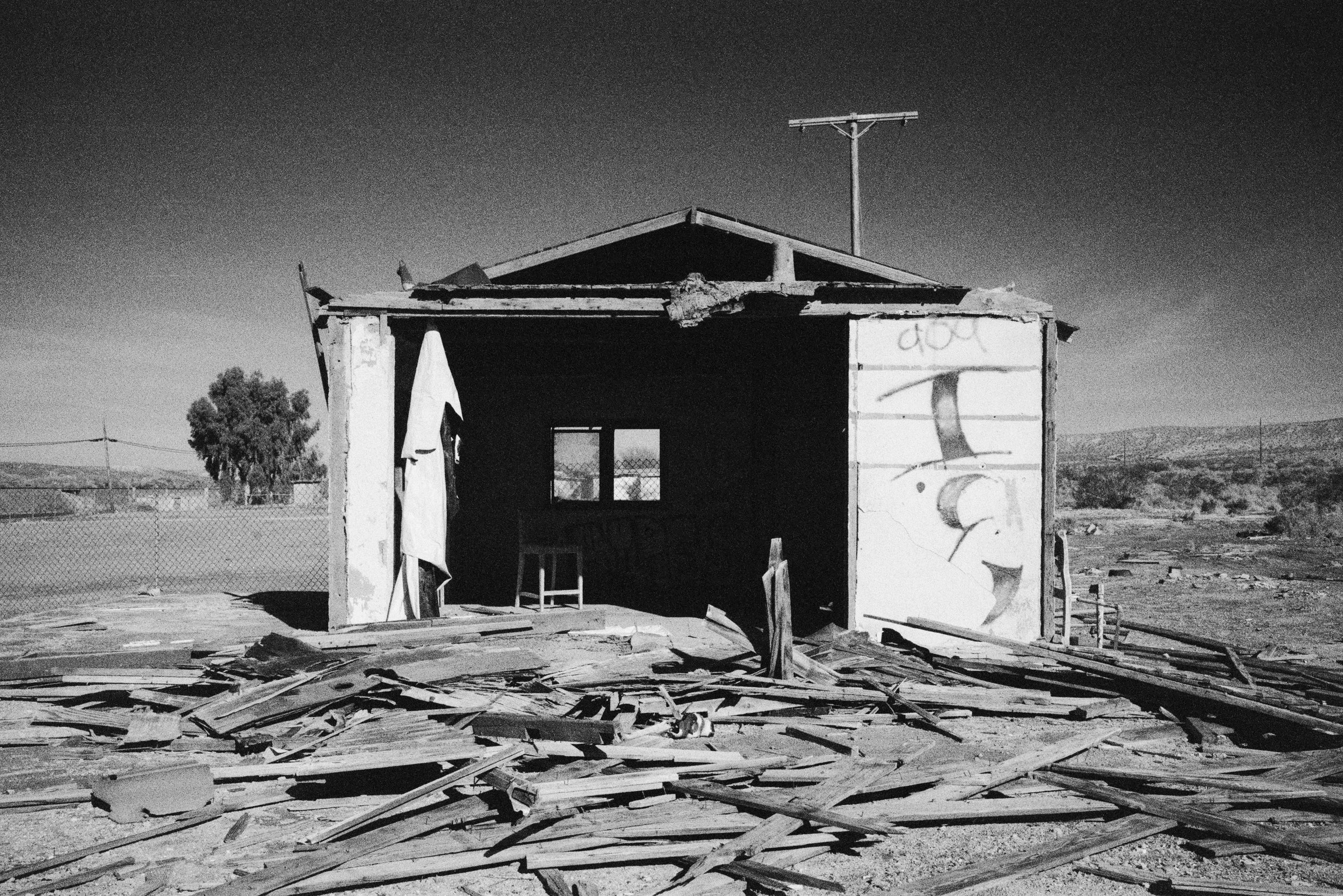 Black and white photo of an abandoned, dilapidated house with debris and broken wooden planks in the foreground. The house has a partially missing roof, an open doorway, and graffiti on the siding. There is a fence and open land in the background.