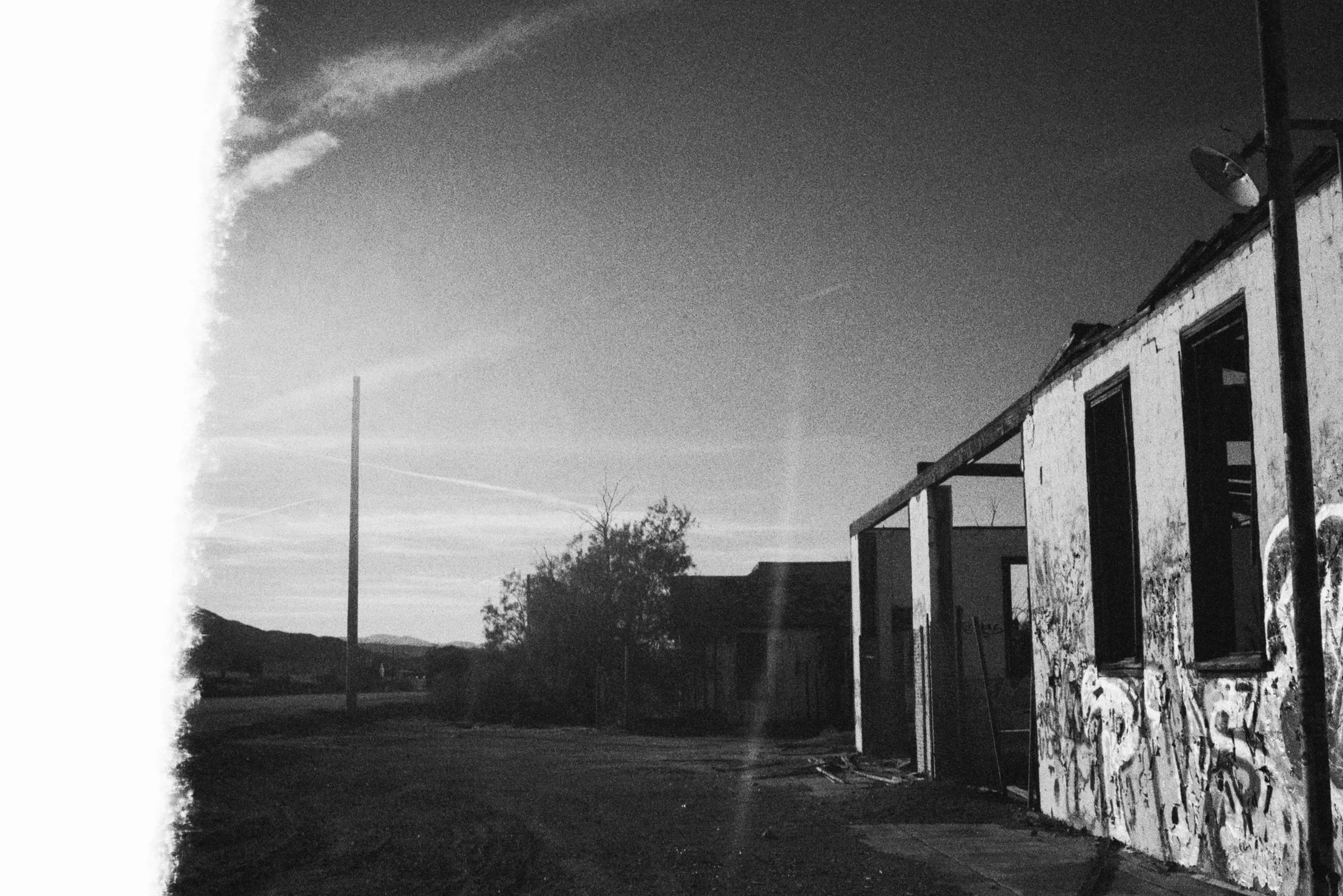 Black and white photo of a rundown building with graffiti, a lit street lamp, trees, power lines, and distant mountains under a sky with trickling clouds.