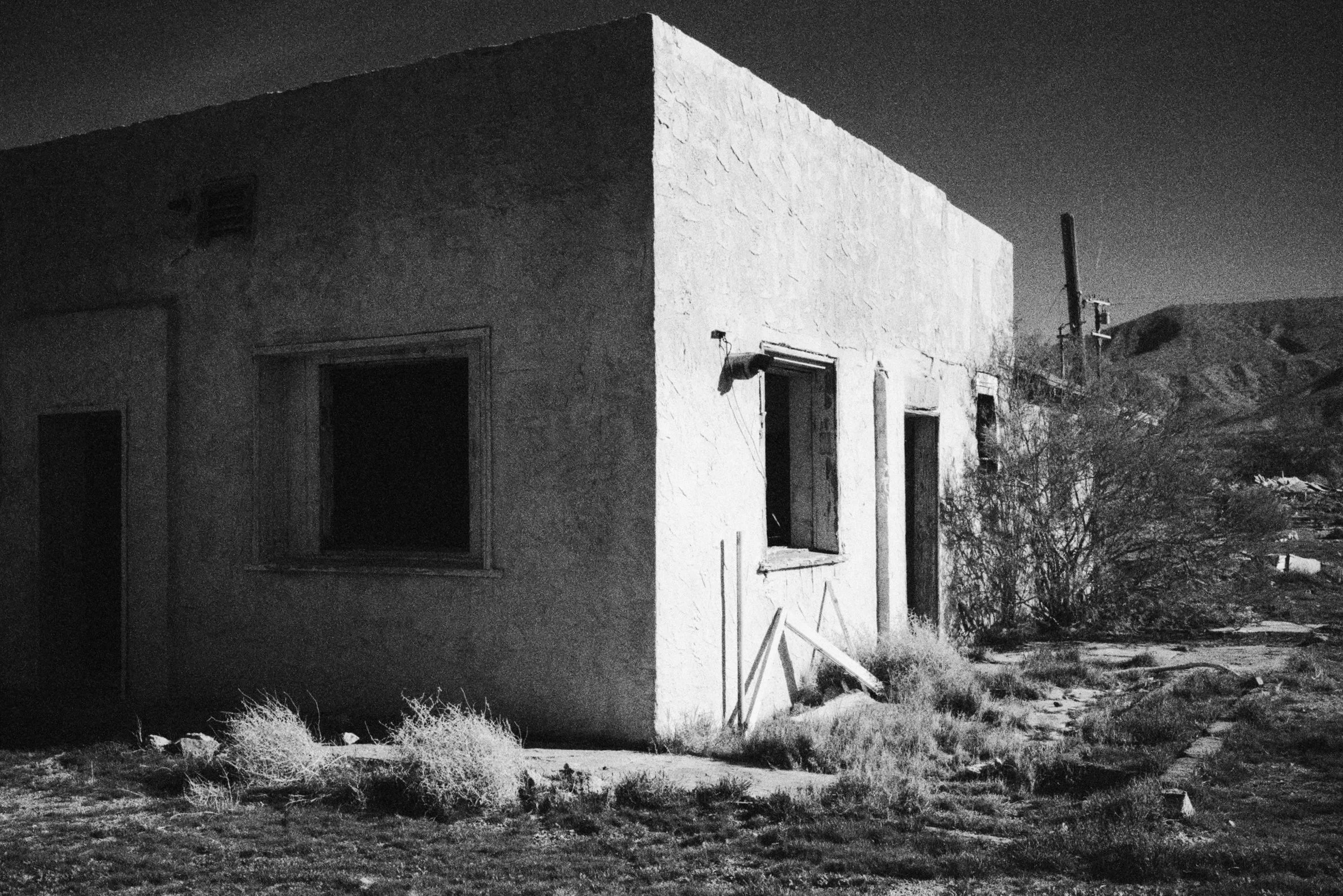 A black and white photograph of an abandoned building in a desert landscape, with overgrown bushes and a rugged terrain. The building has a flat roof, boarded-up windows, and an exterior light fixture. There is a utility pole with wires in the backgr