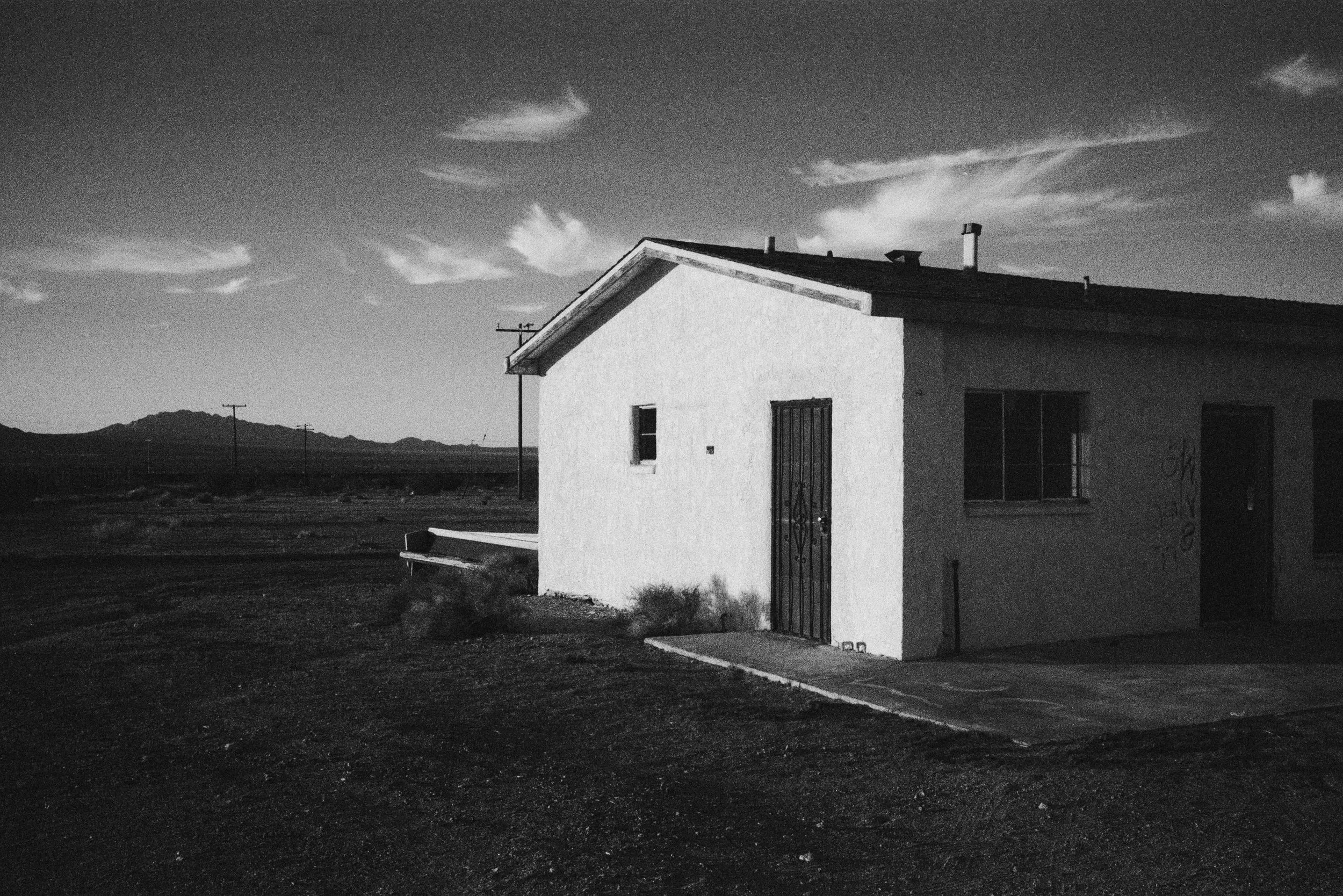 A small, white house with a metal gate and a small window, set in a desert landscape with mountains in the background and a cloudy sky.