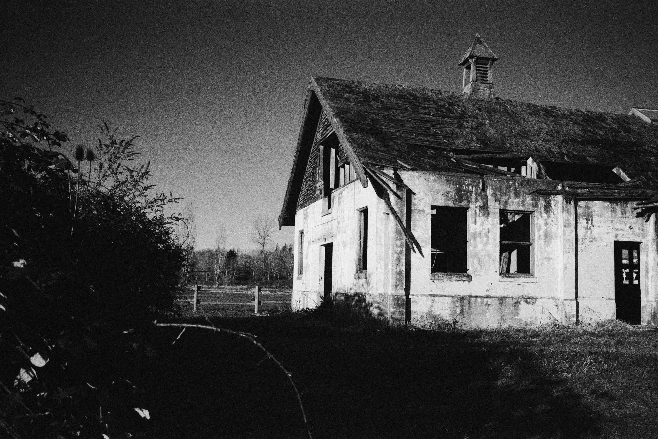 Black and white photo of an abandoned, dilapidated house with broken windows and a damaged roof, surrounded by an open field and sparse trees in the background.