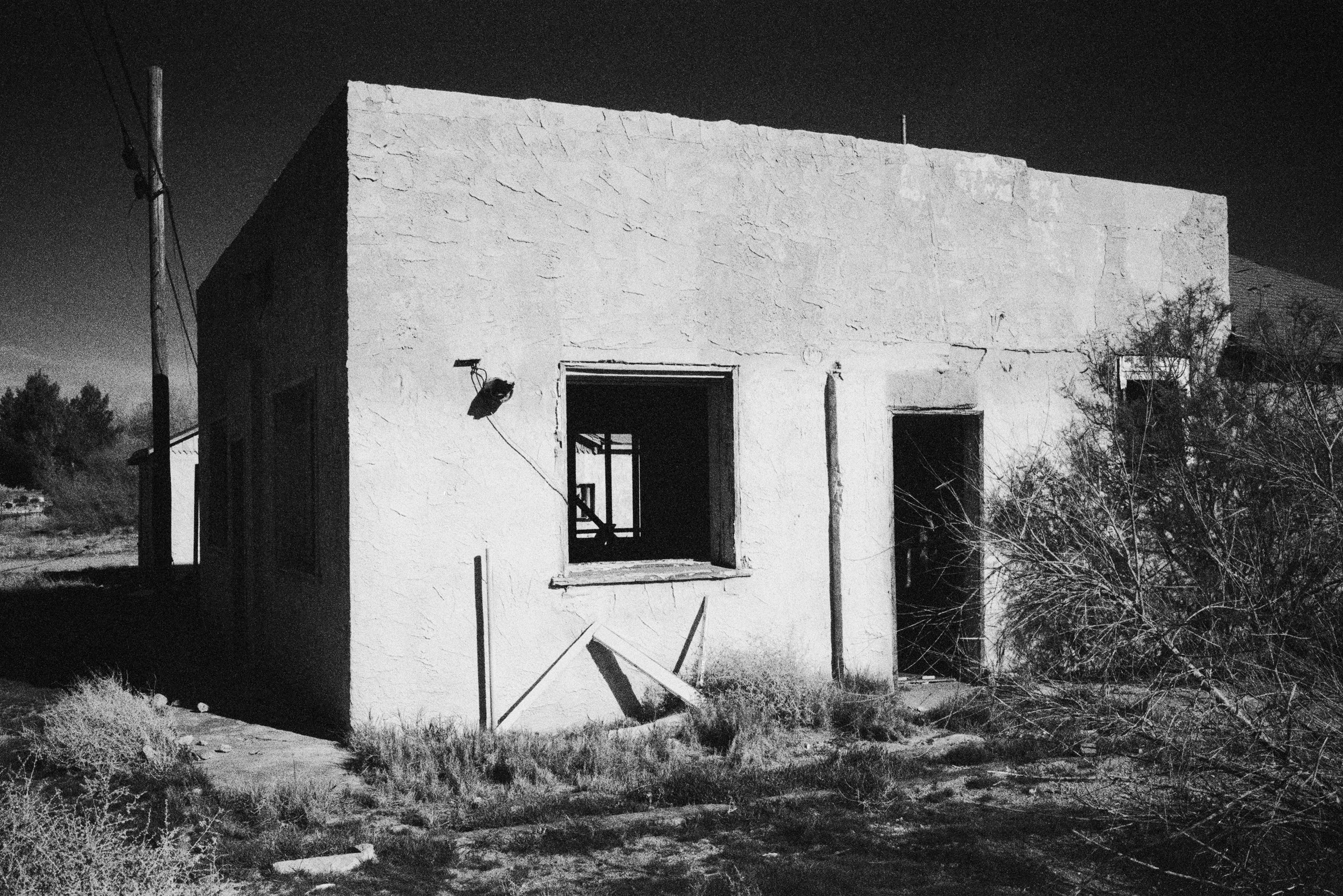 An abandoned, weathered building with missing windows, surrounded by dry bushes and grass, under a dark sky.