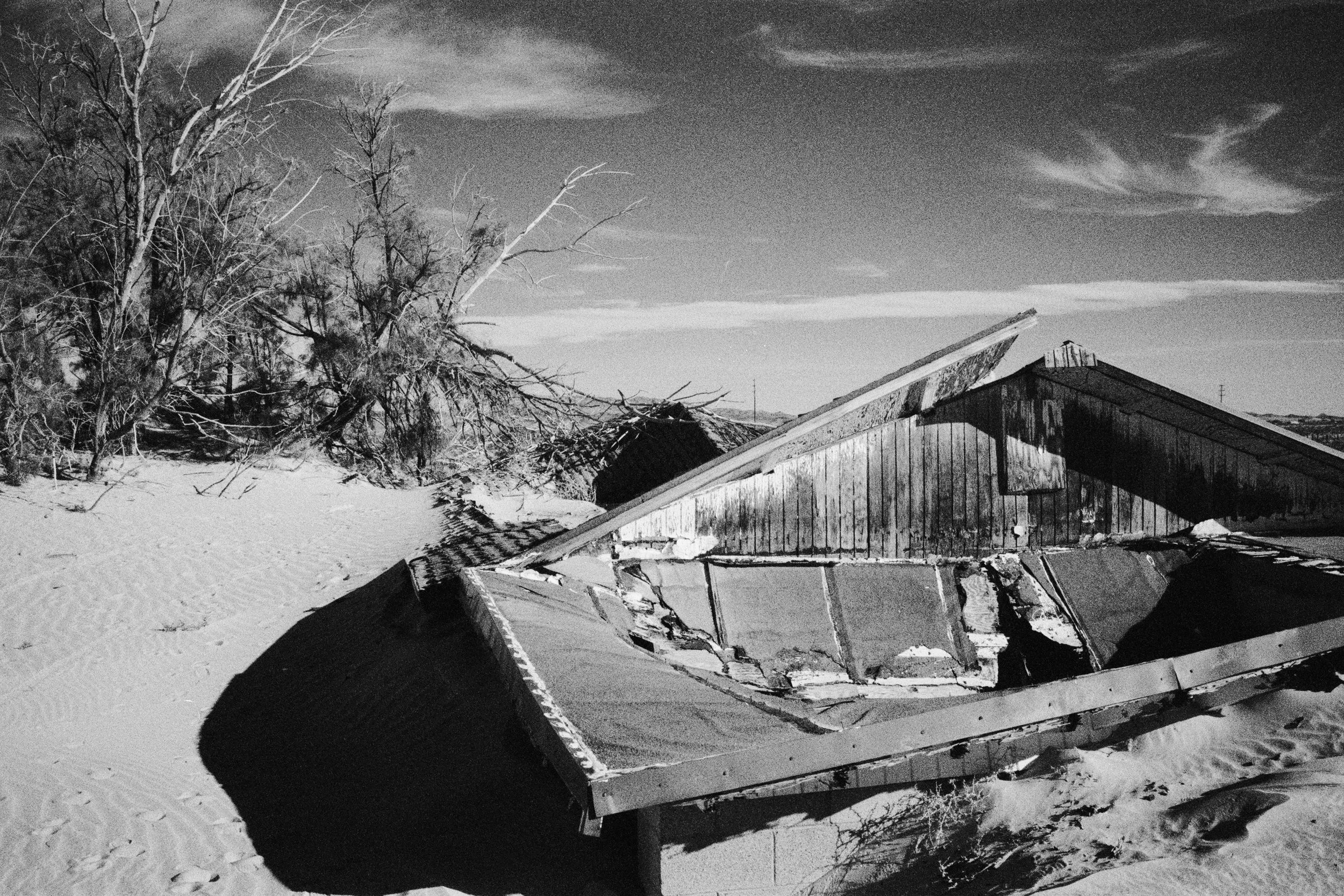 A black and white photo of a toppled billboard sign on a snowy landscape, with a wooden building behind it and trees in the background.