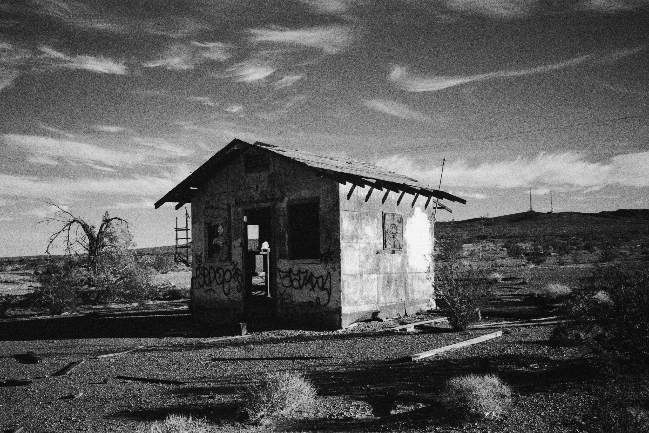 An abandoned, graffiti-covered house in a desert landscape, with sparse vegetation and hills in the background, under a cloudy sky.