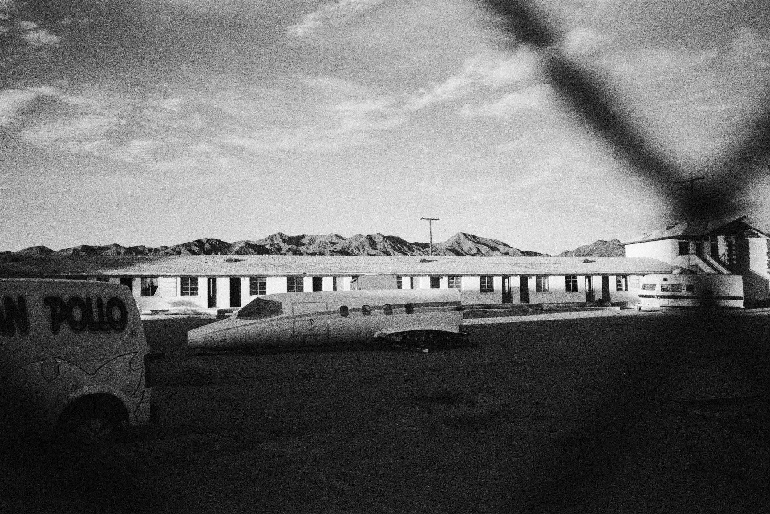 A black and white photo of a desert landscape with mountains in the background. There is a single-story building with several windows in the middle of the scene, and a sci-fi style aircraft or spaceship parked on the ground nearby. Part of a van with