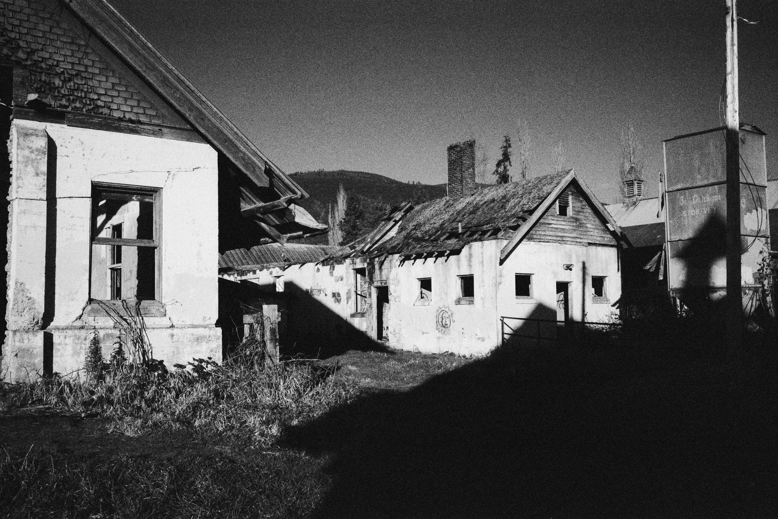Black and white photo of abandoned, weathered houses with deteriorating roofs, broken windows, and overgrown plants in front. Mountainous landscape in the background and utility pole on the right.