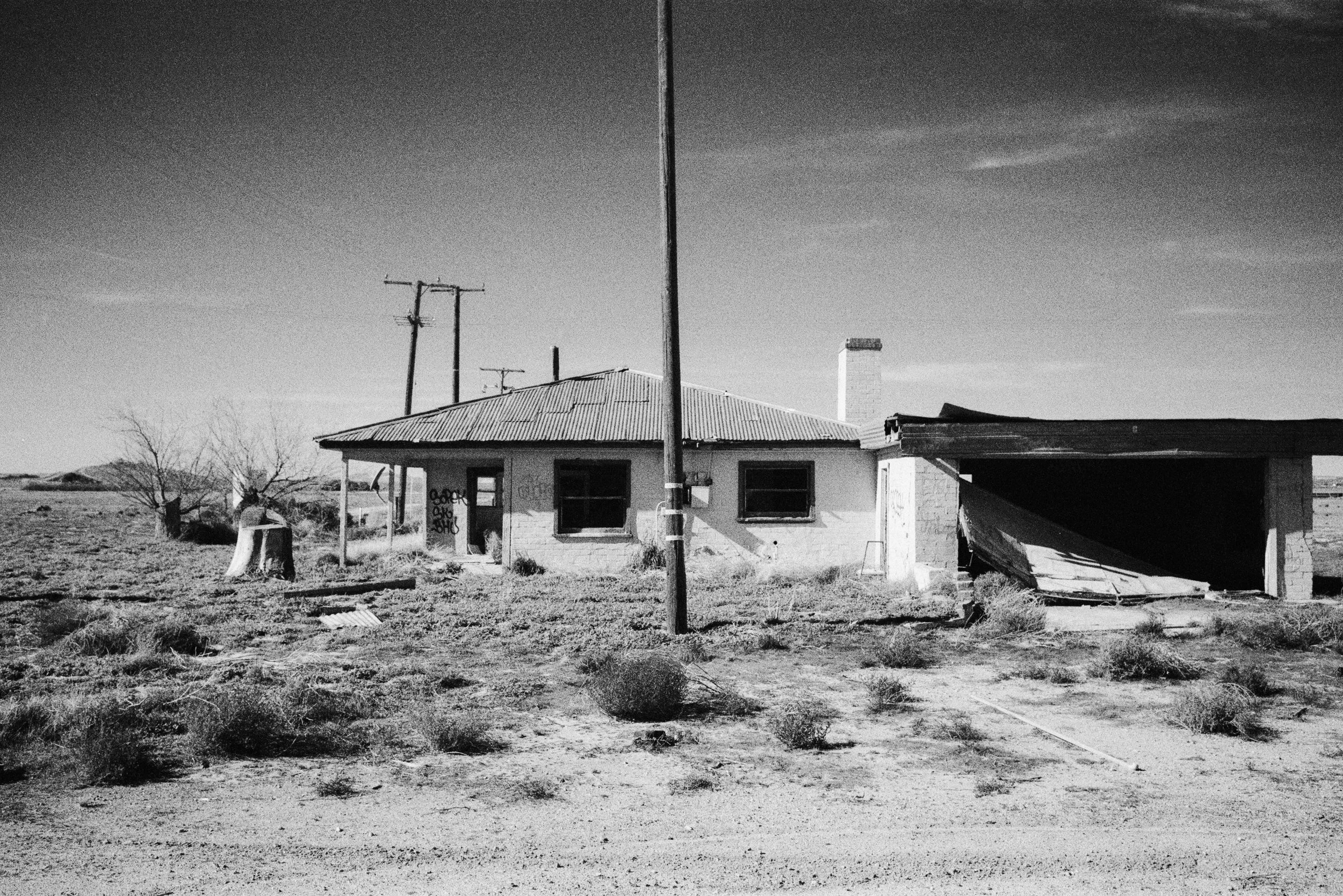 Black and white photo of an abandoned, dilapidated house in a barren landscape with dry bushes, an old tree stump, and power lines under a clear sky.