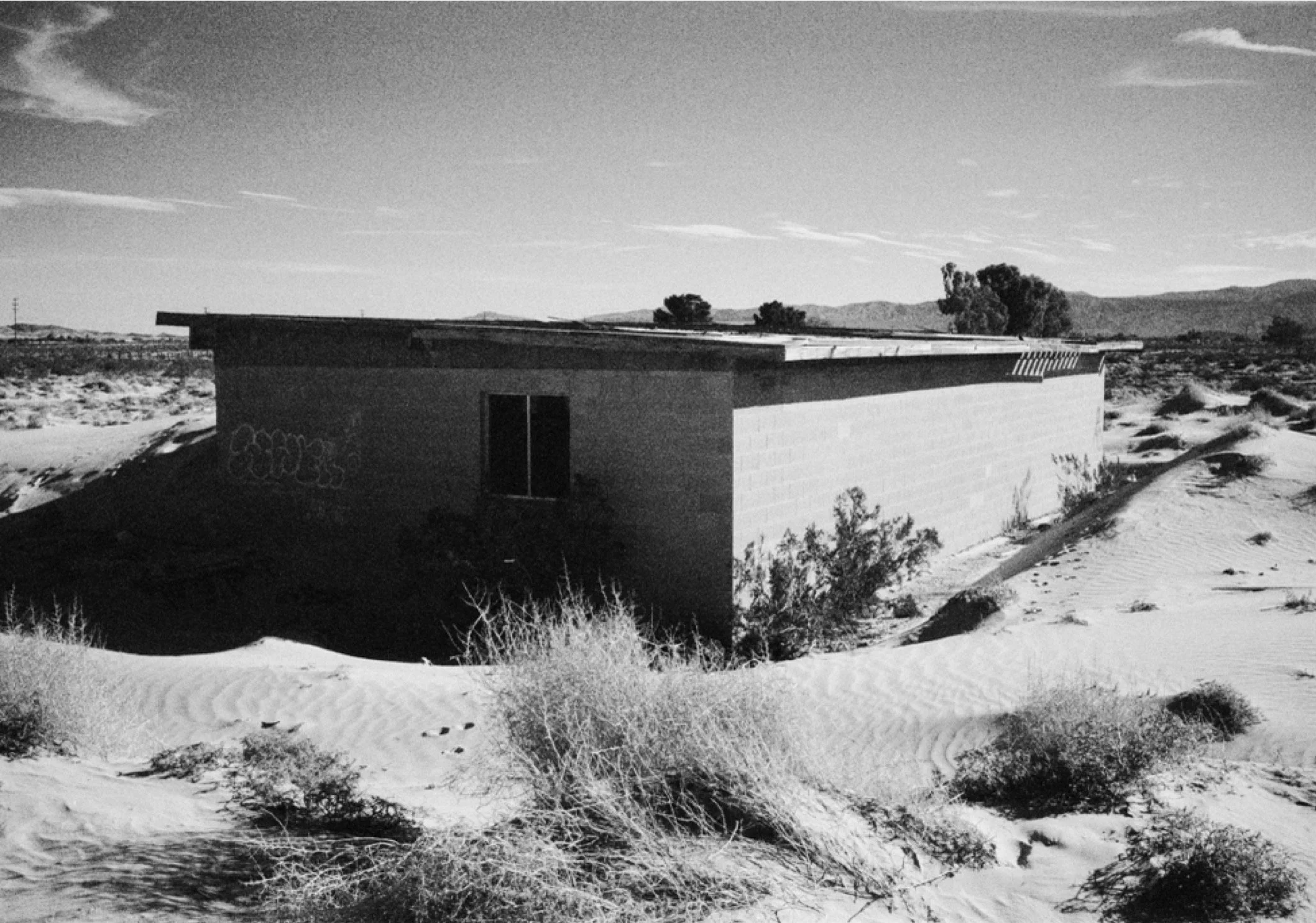 A small, flat-roofed building in a desert landscape with sand dunes and sparse shrubs, under a partly cloudy sky.