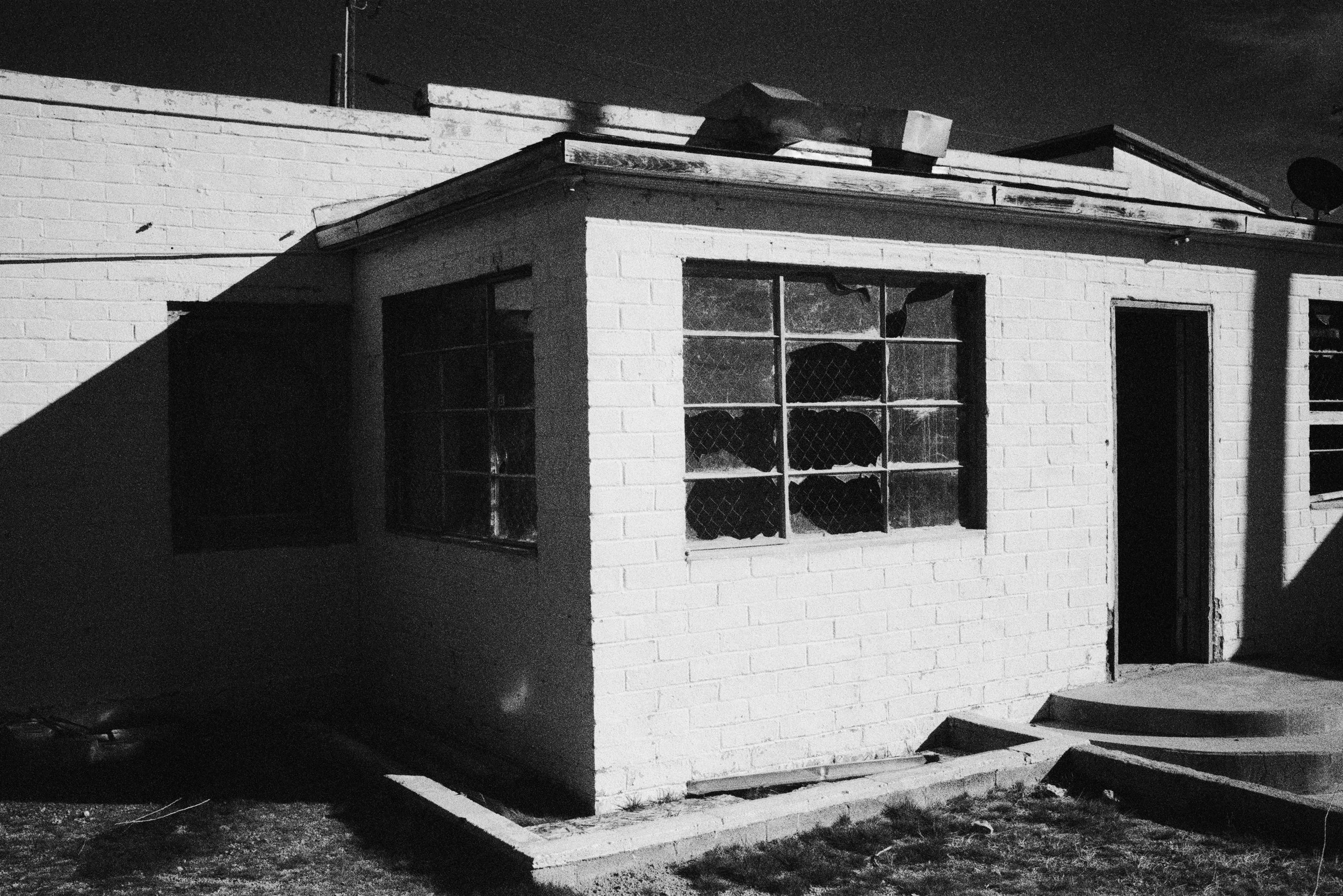 A black and white photo of a small brick building with a flat roof, windows covered with wire mesh, and a door, with shadows cast on the wall.