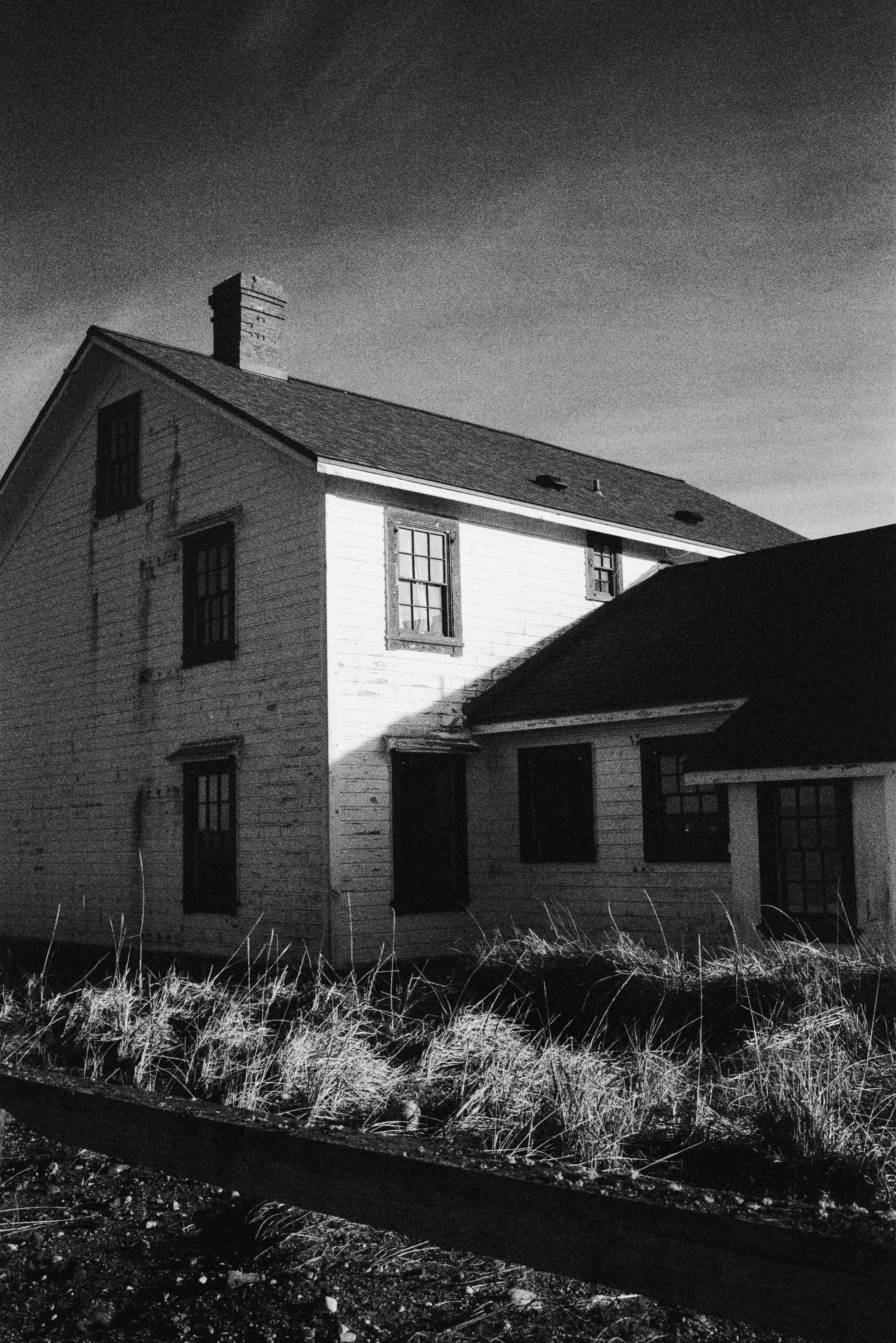 A black-and-white photo of an old, two-story house with a chimney, boarded-up windows, and weathered exterior. Overgrown grass in the foreground and a cloudy sky in the background.