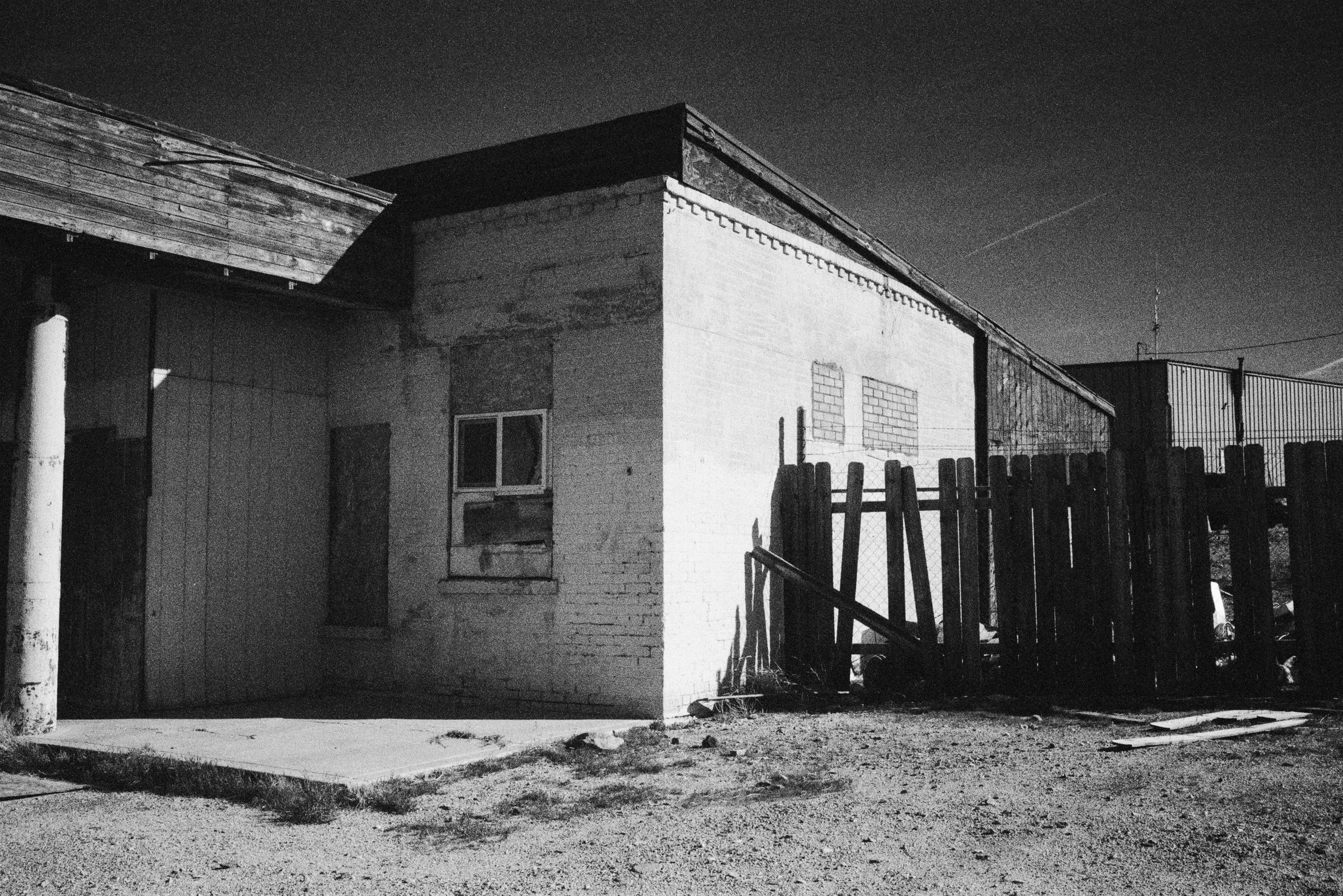 Black and white photo of a rundown building with boarded-up window and a makeshift fence, under a dark night sky.
