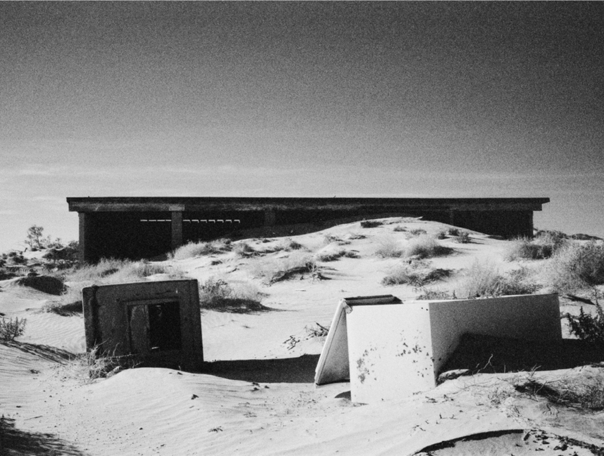 Desolate sand dunes in a desert with a dilapidated structure and debris, black and white photography.