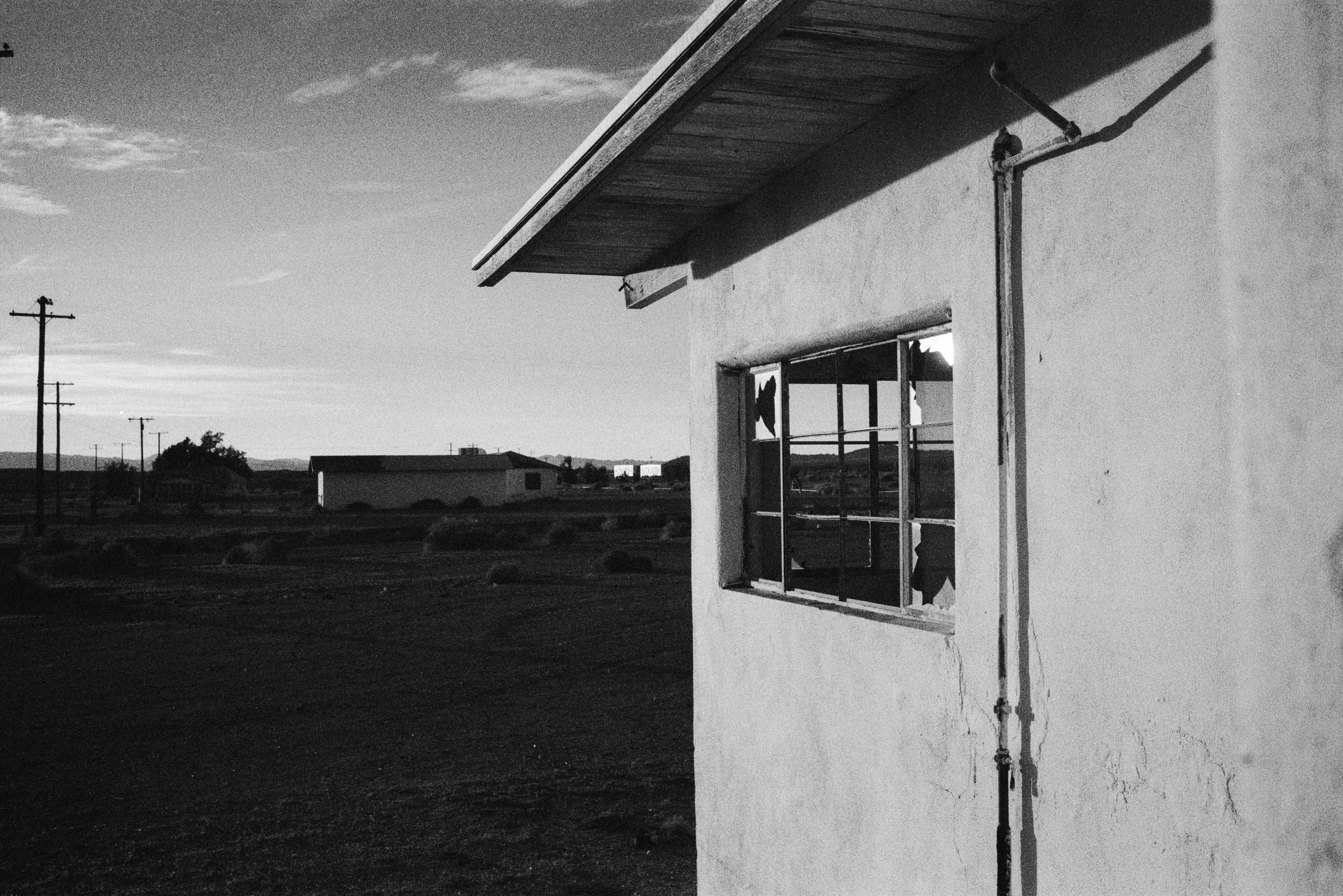 A black-and-white photo of a house with a broken window and exposed piping, set in a rural landscape under a cloudy sky.
