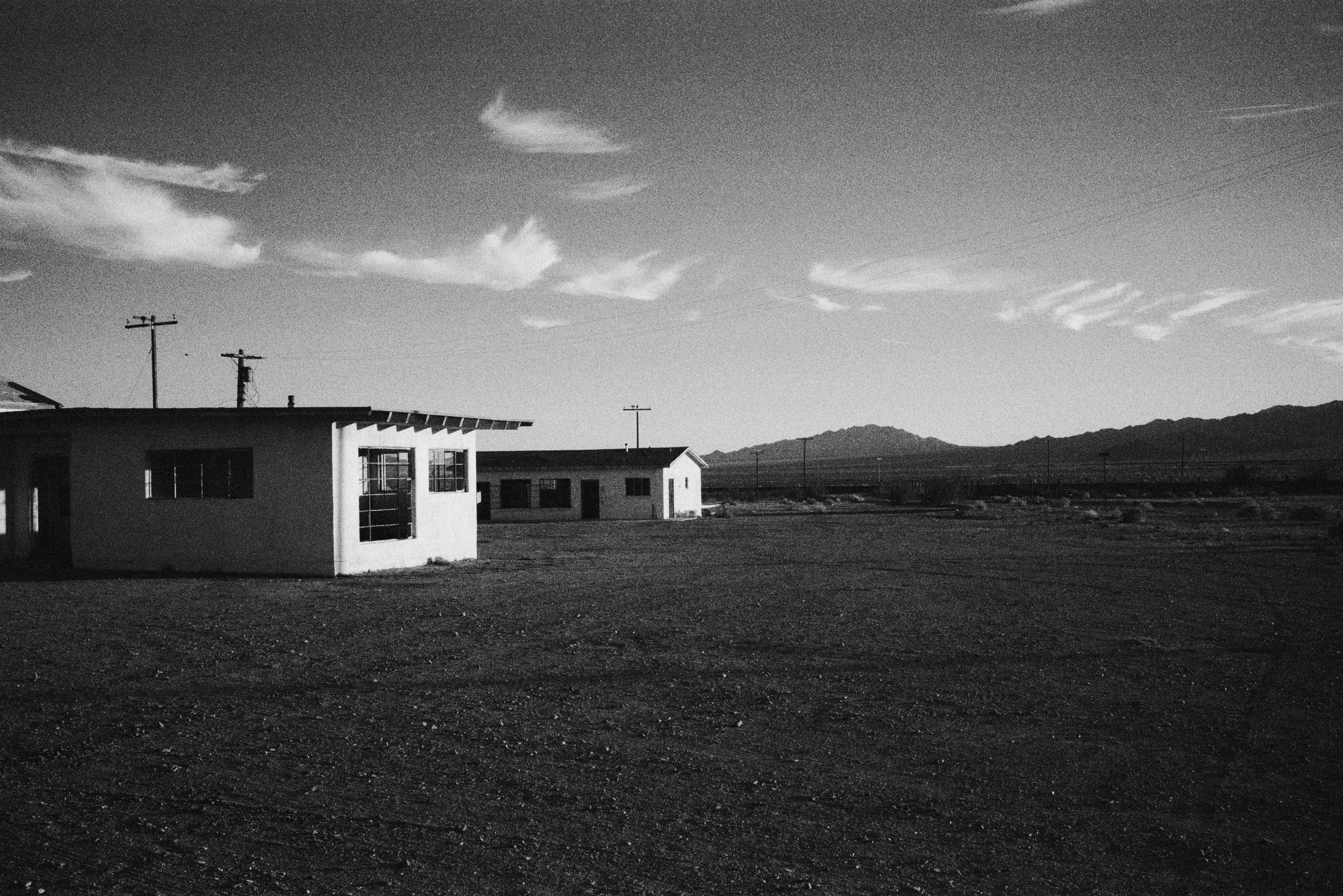 A black and white photo of a remote, barren landscape with a few small, simple buildings and utility poles, mountains in the distance, and a partly cloudy sky.
